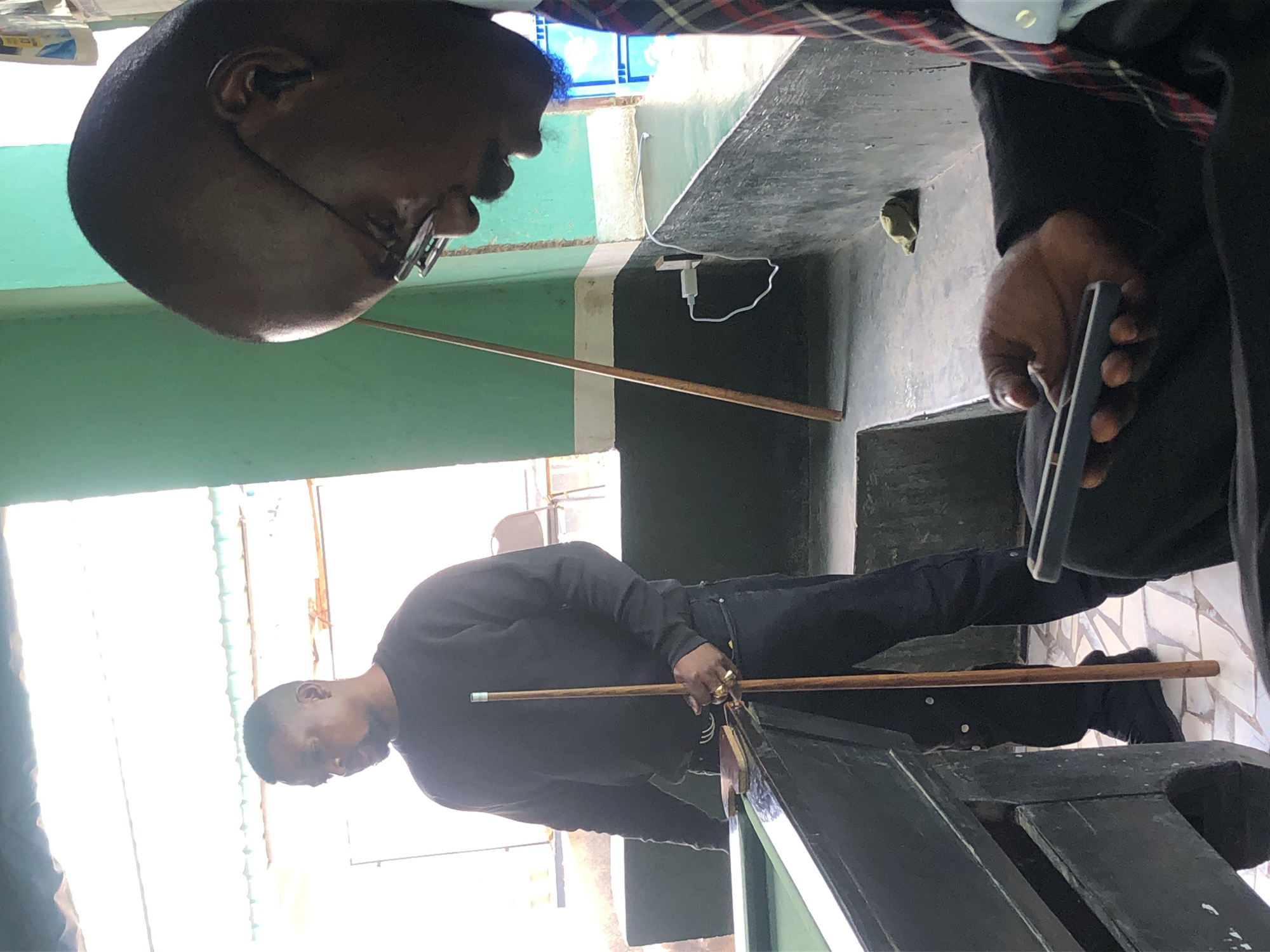The image depicts two Black men in an indoor setting, possibly a recreational facility or bar, with a pool table in the foreground. The man on the left, wearing a dark long-sleeved shirt and dark pants, stands holding a pool cue vertically. He has short hair and is looking down. The man on the right, in the foreground, is wearing glasses and a suit jacket with a plaid tie. He is holding a smartphone in his left hand and looking down at it, his head partially in profile. The environment includes a pool table with a green felt surface and dark wooden edges. A light-colored ceiling with visible beams is present, and a green wall is visible in the background. A white electrical outlet with a charging cable is plugged into the wall. The floor appears to be tiled.