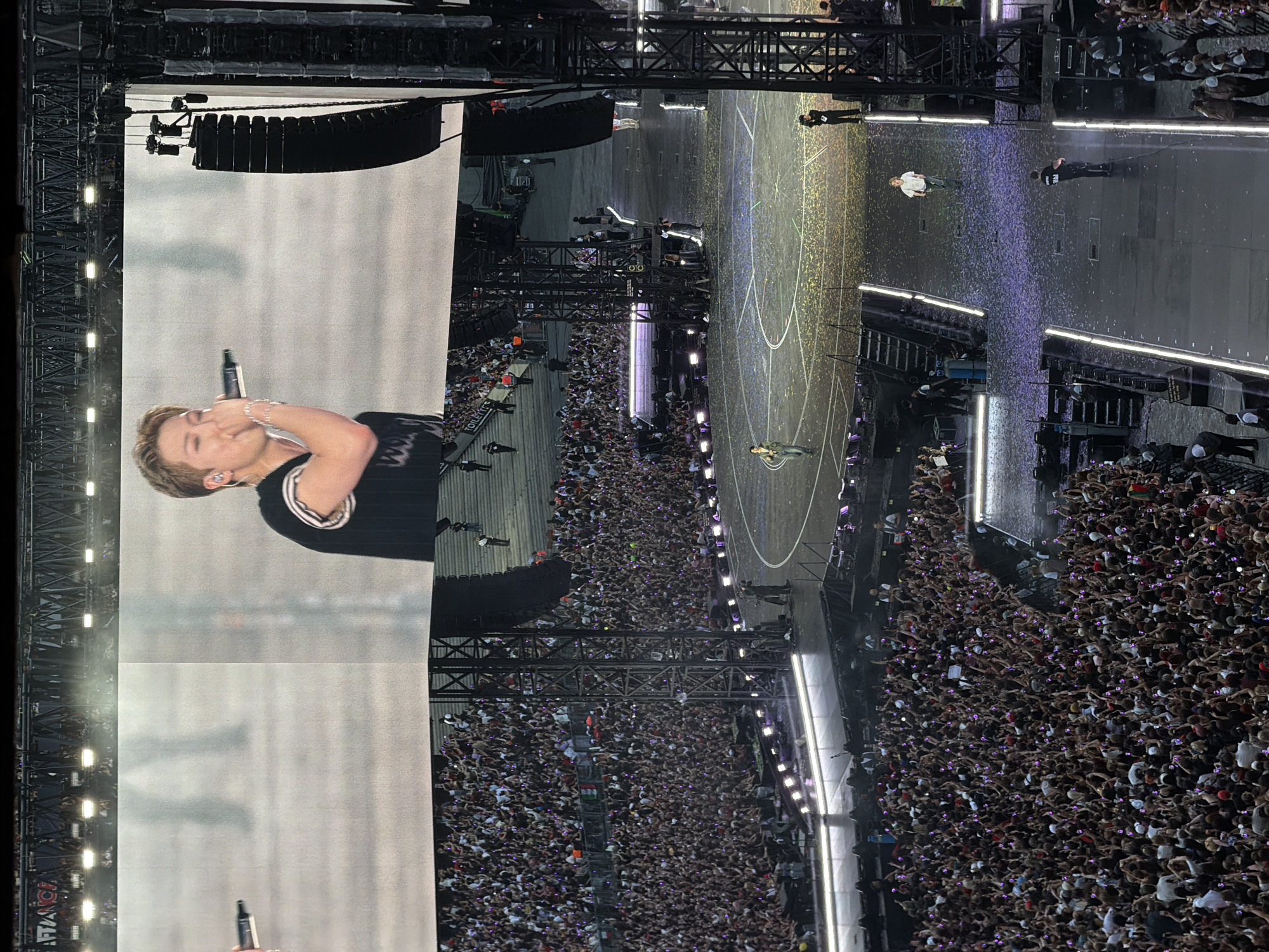 The image depicts a large-scale outdoor concert. In the background, a massive LED screen shows a close-up of a male performer singing into a microphone. He has blonde hair and is wearing a dark vest over a white shirt with striped trim. Below the screen, a vast crowd of concertgoers fills tiered seating and stands, many holding up illuminated phones, creating a sea of lights. The concert stage is visible in the foreground, with several performers walking across it. Confetti is scattered across the stage floor. The overall environment suggests a dynamic and energetic live music event in a large, open-air venue.
