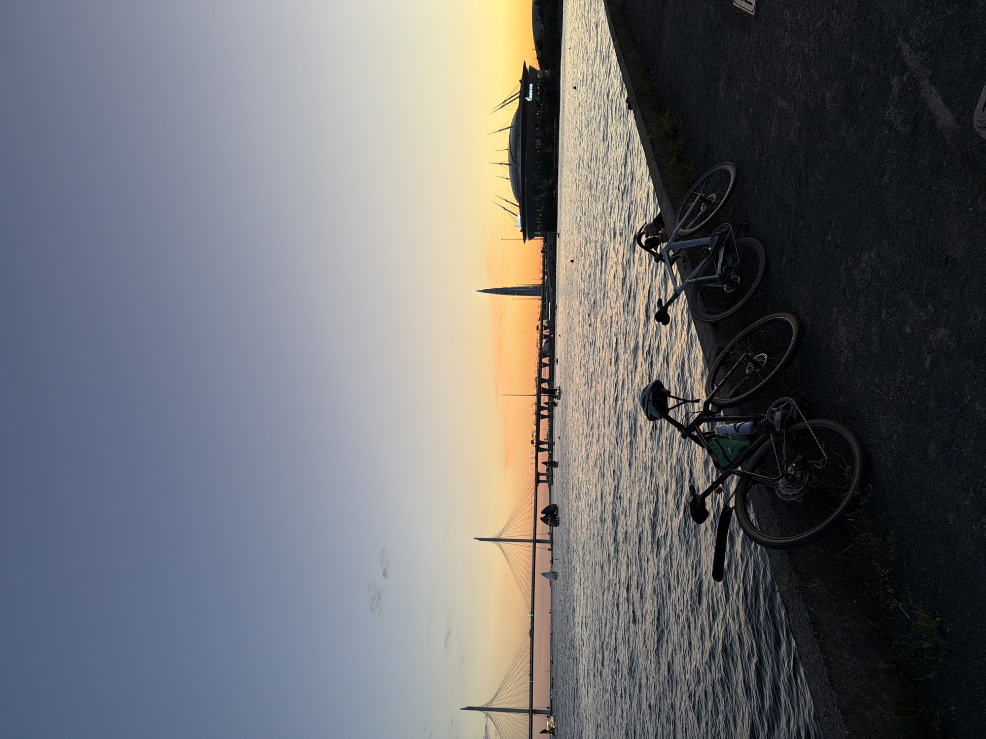 This image captures a sunset scene in Saint Petersburg, Russia, likely near the Gulf of Finland. In the foreground, three bicycles are parked along a concrete embankment, leaning against each other. One bicycle, a darker color, has a green bag and a water bottle attached to its frame, and a helmet rests on its handlebars. Two other bicycles, lighter in color, are positioned next to it. The water of the bay is visible, with gentle waves reflecting the warm hues of the sunset. In the background, a large, modern bridge stretches across the water, with distinctive cable-stayed towers. Further in the distance, a tall, slender skyscraper and a domed stadium with a distinctive roofline are visible against the colorful sky. Several sailboats are dotted on the water, adding to the serene atmosphere. The sky displays a gradient of colors, transitioning from a pale blue at the top to warm oranges and yellows near the horizon.