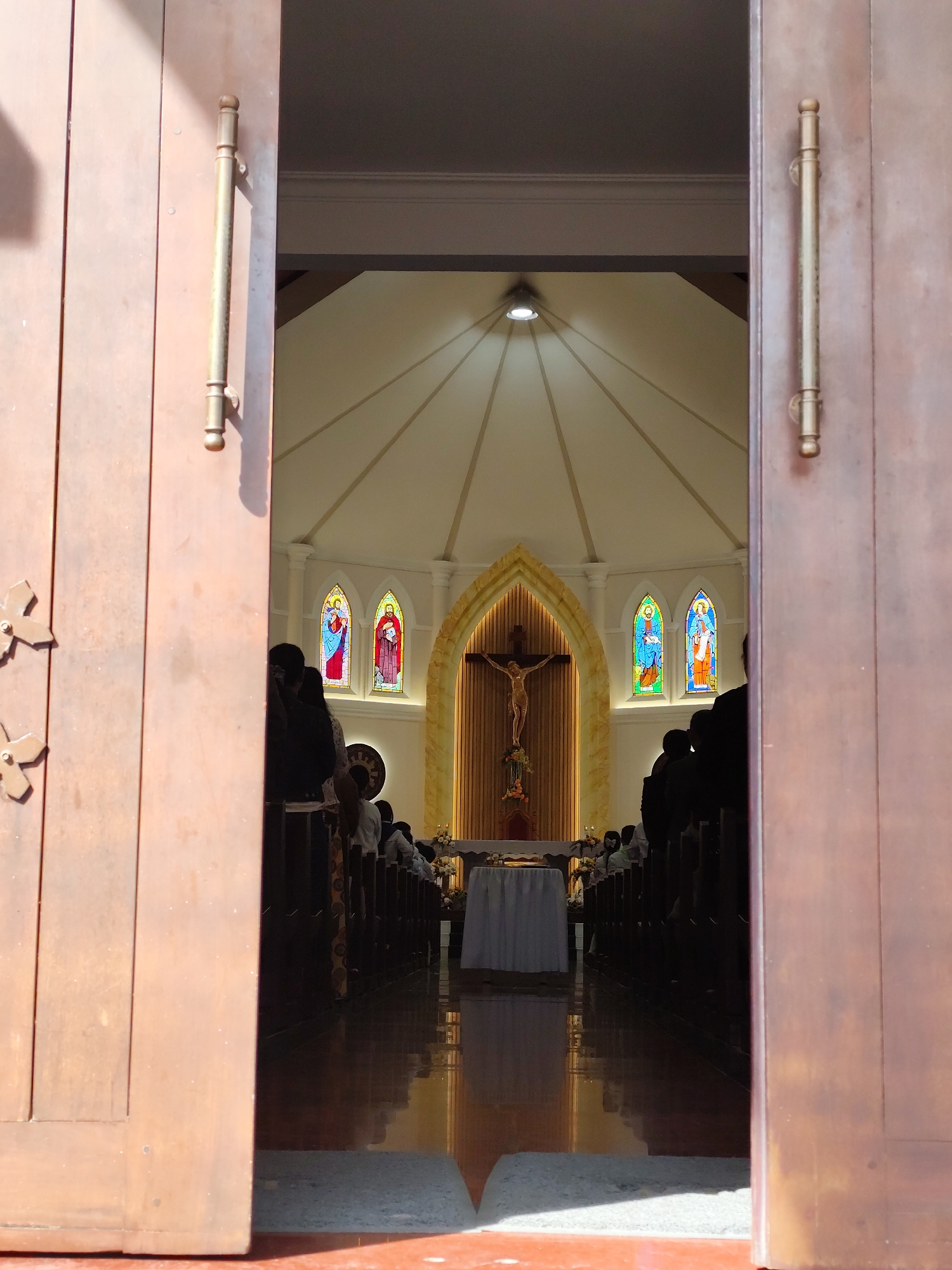 The image depicts the interior of a church in Lâm Viên - Đà Lạt, Vietnam, as viewed through a partially open set of wooden doors. The church features a high, vaulted ceiling and stained-glass windows. Rows of pews are visible on either side of the aisle, with several people seated. A crucifix hangs above the altar, which is covered with a white cloth. The polished floor reflects the interior lighting. The door frames are adorned with ornate handles and decorative elements.