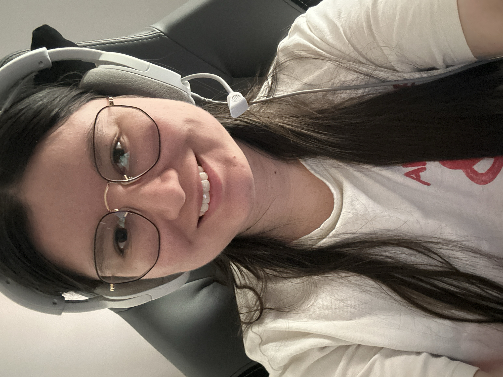 A close-up selfie shows a person with light skin, brown hair, and glasses, smiling. They are wearing a white t-shirt and grey headphones with a microphone. The person is sitting in a dark grey chair. The background is a plain white wall. The image was not taken in Câmara de Lobos, Portugal as there are no elements in the picture that would support this claim.