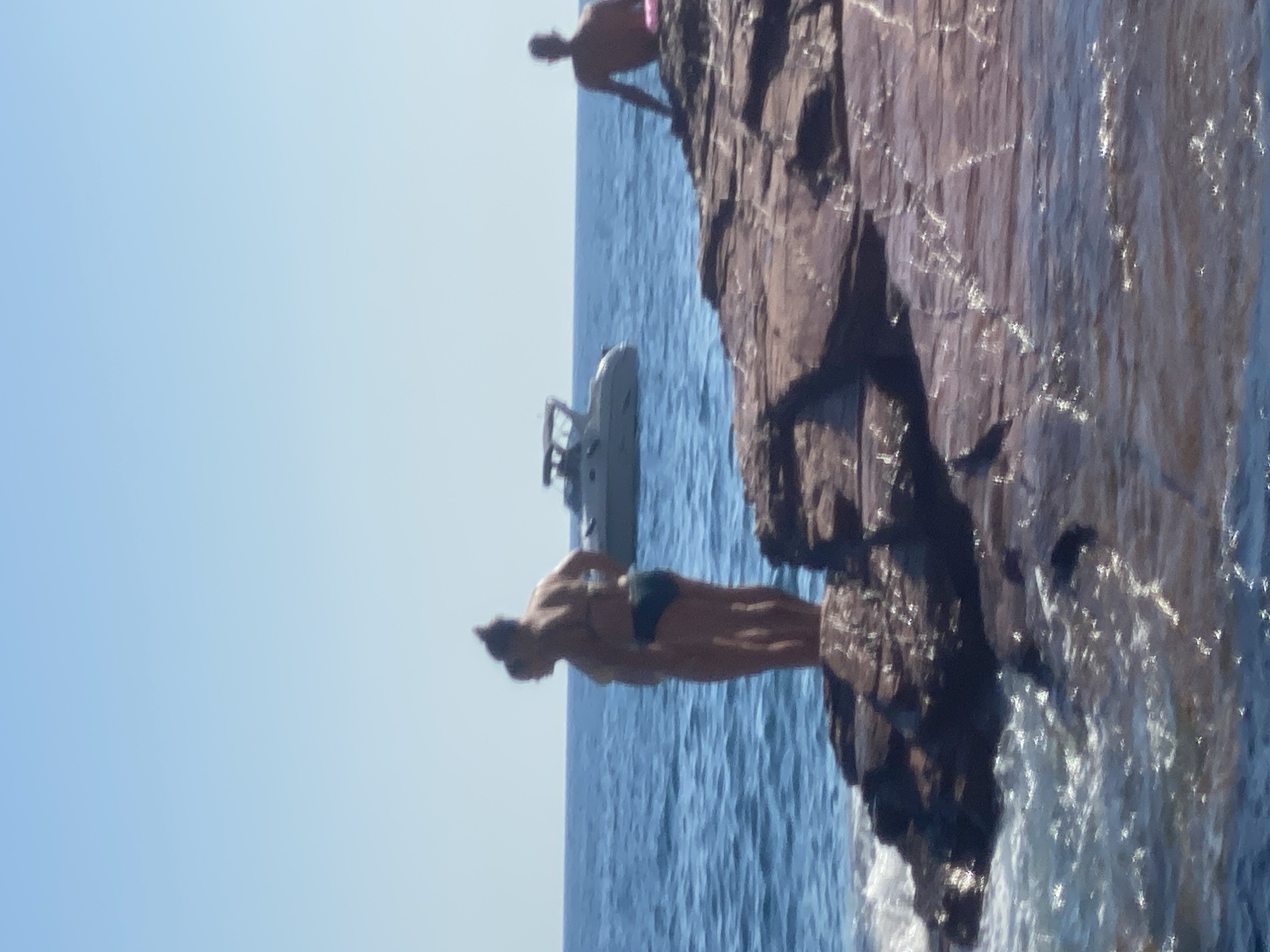 The image shows a clear blue sky above a calm blue sea. In the foreground, reddish-brown rocks jut out into the water. On the rocks, two people are standing near the edge. The person on the left is facing away from the camera, while the person on the right is partially visible. Further out in the water, a white boat is moving across the scene. To the right of the boat, another person is sitting on the rocks, facing away from the camera. The overall scene appears to be a sunny day at the coast, possibly in Sutomore, Montenegro, given the context.