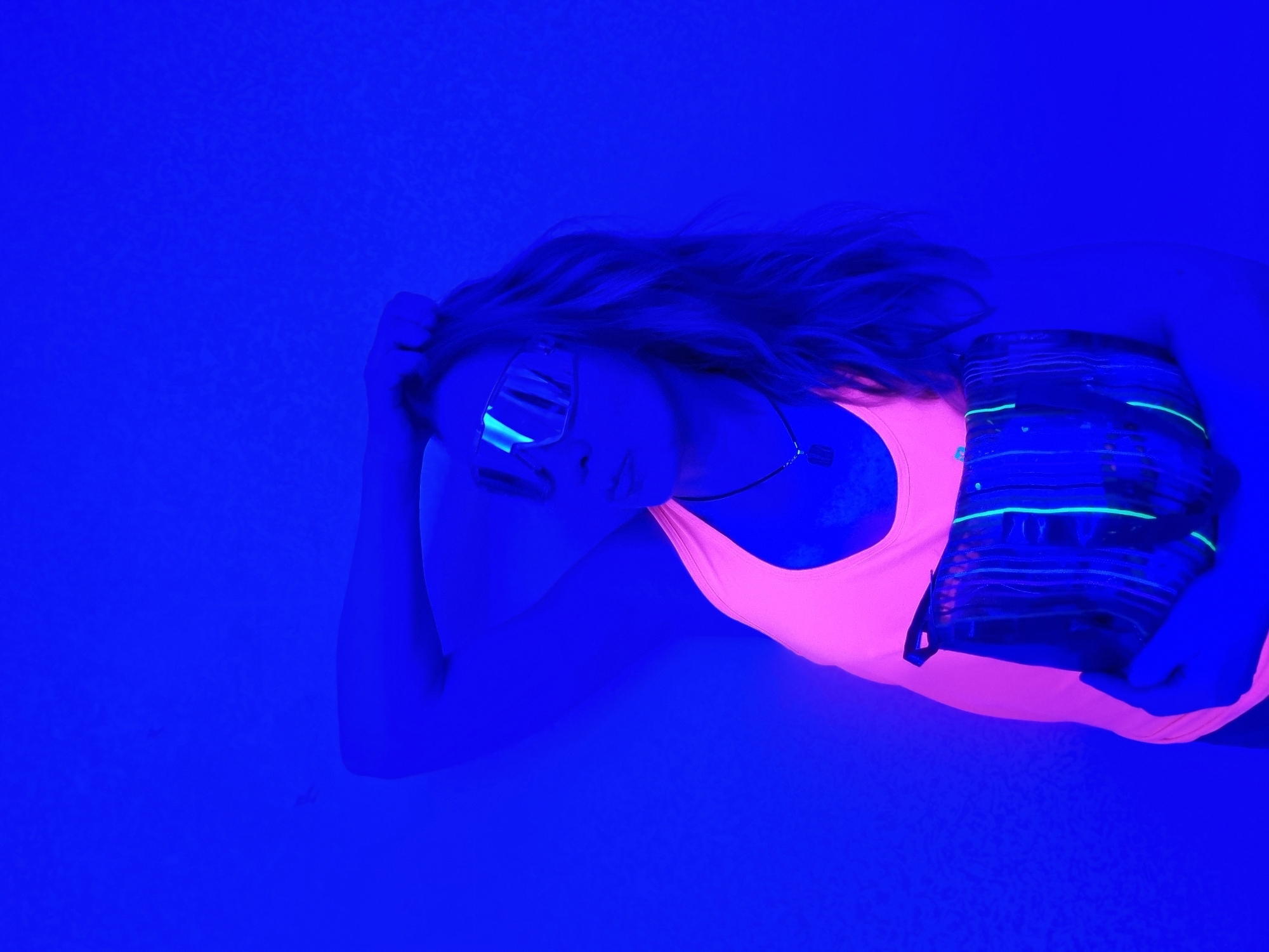 A woman is lying on a flat surface under blue light. She has blonde hair and is wearing mirrored sunglasses, a pink tank top, and a necklace. She is holding a striped object in her arms. The background is a uniform blue. There is no visible information to suggest this image was taken in Chaikovsky, Russia.