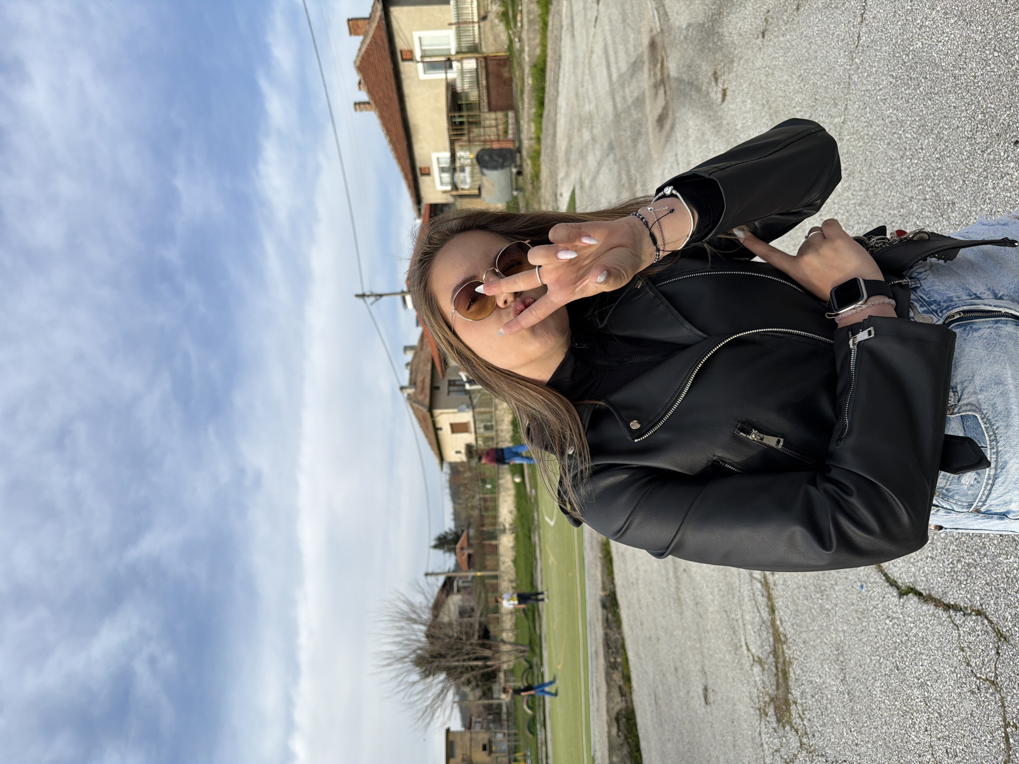 A woman in a black leather jacket, sunglasses, and blue jeans poses with a peace sign in front of an outdoor recreational space. This space, identified as being in the city of Popintsi, Bulgaria, features a green field with white lines (potentially a soccer field), surrounded by a concrete walkway and low fencing. In the background, there are houses with visible roofs, trees, and other people on the field. The sky is partly cloudy. The ground in the foreground is cracked asphalt. The woman wears an Apple watch and bracelets on her right wrist. The buildings in the background suggest a residential area.