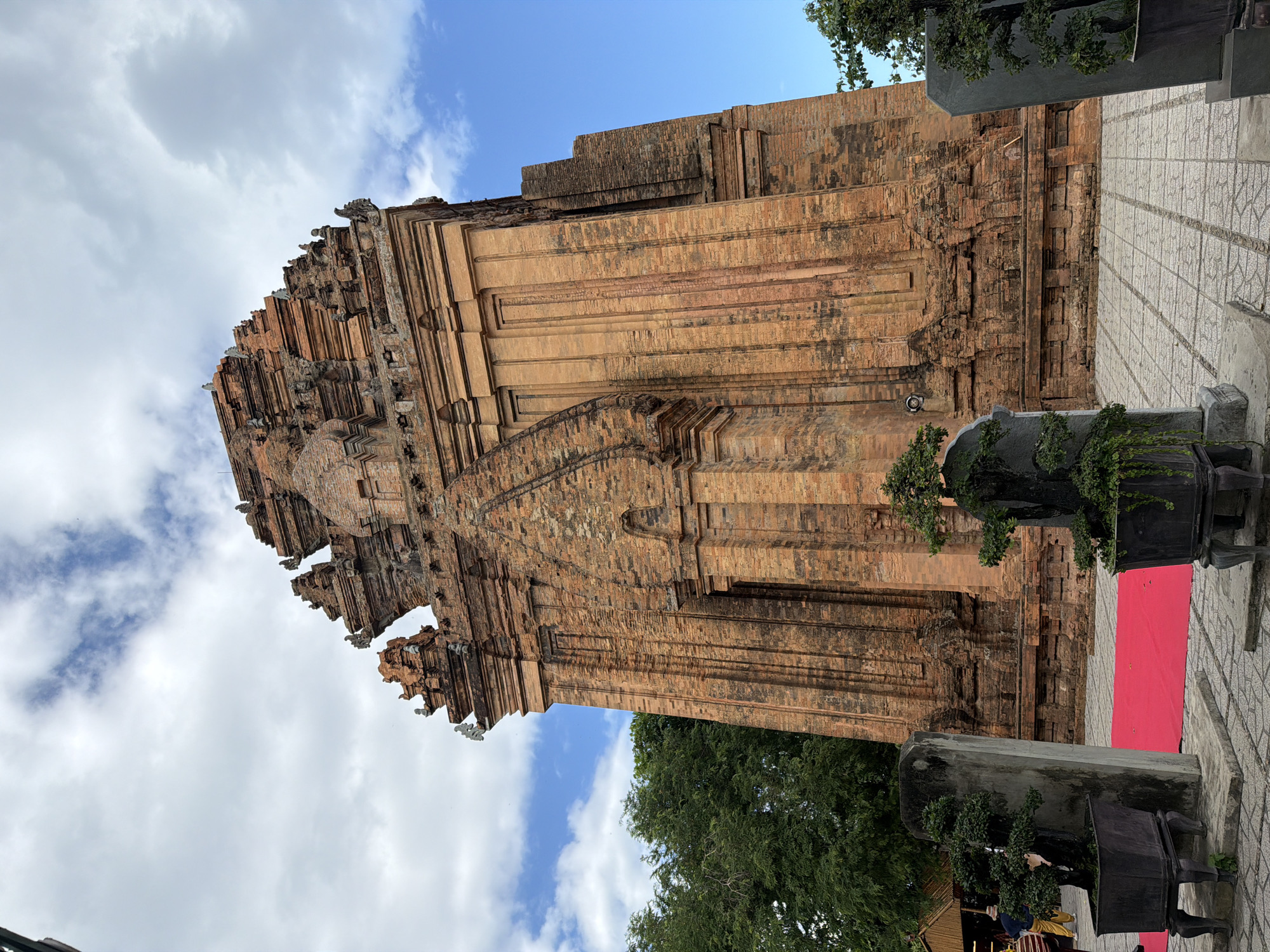 The image displays the Po Nagar Cham Towers, a complex of temples located in Nha Trang, Vietnam. The primary subject is the central, largest tower, constructed from reddish-brown brick and featuring intricate architectural details, including tiered roofs and decorative carvings. The tower is a significant example of Cham architecture. In the foreground, a vibrant red carpet is laid out on a paved courtyard, flanked by stone pillars and planters containing bonsai trees. To the left, a portion of a wooden structure, possibly a stall or pavilion, is visible, with two individuals in casual attire walking in the background. Lush green trees frame the right side of the towers. The sky above is partly cloudy, with patches of blue sky visible. The overall scene suggests a cultural or tourist site, with elements of both ancient architecture and modern amenities.