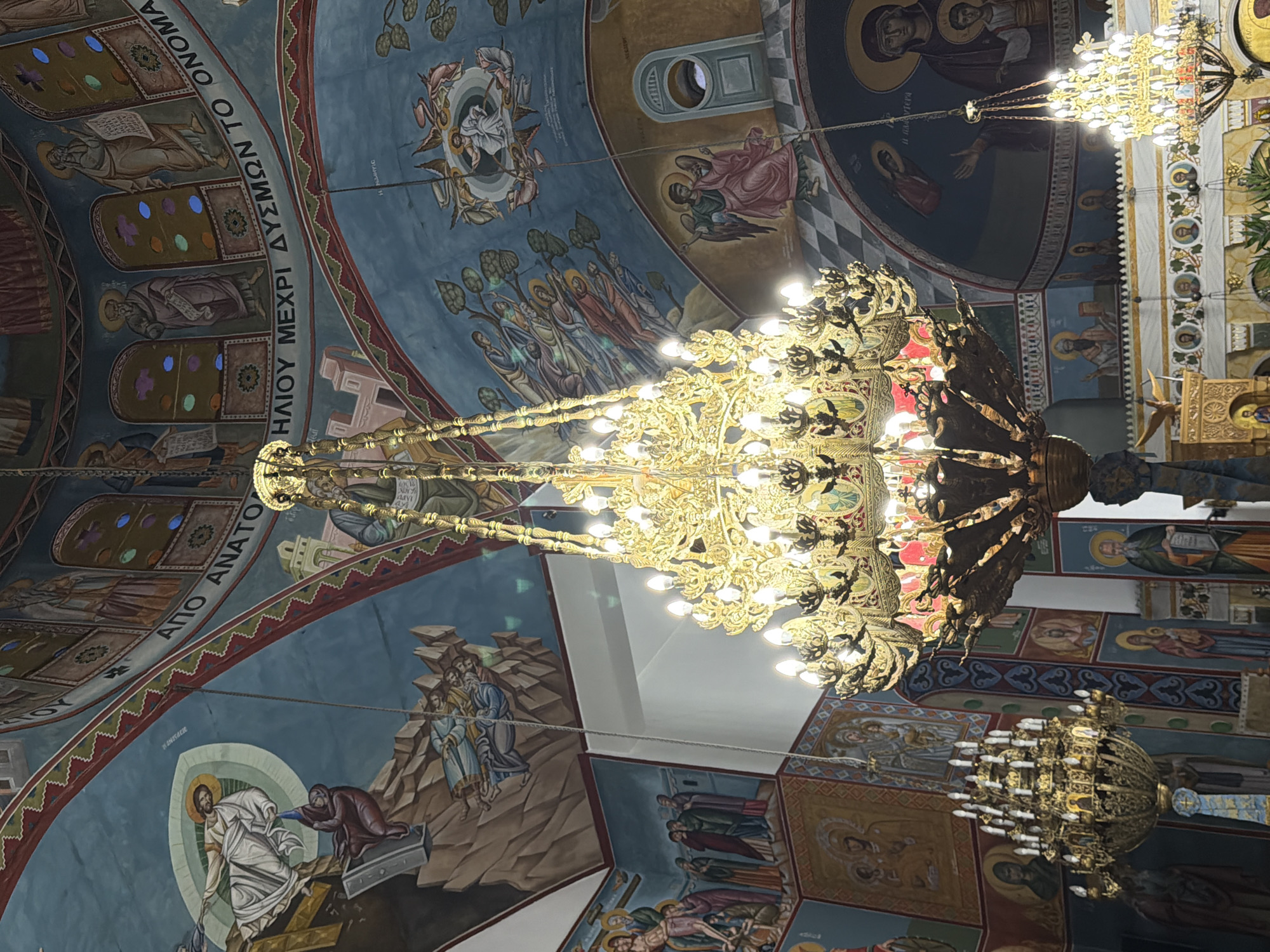 The image shows the interior of a richly decorated church, likely an Orthodox Christian church, given the iconography. The ceiling is adorned with vibrant frescoes depicting religious scenes and figures, including Jesus and various saints. Several ornate, multi-tiered chandeliers, illuminated with many bulbs, hang from the ceiling, casting a warm glow on the scene. The walls are also covered with religious paintings and intricate decorative borders. The overall impression is one of grandeur and spiritual significance.
The location is identified as the Municipal Unit of Alyzia, Greece. Without more specific information or external landmarks, it is impossible to pinpoint the exact church within Alyzia. However, the style of the frescoes and the architecture are consistent with traditional Greek Orthodox churches.