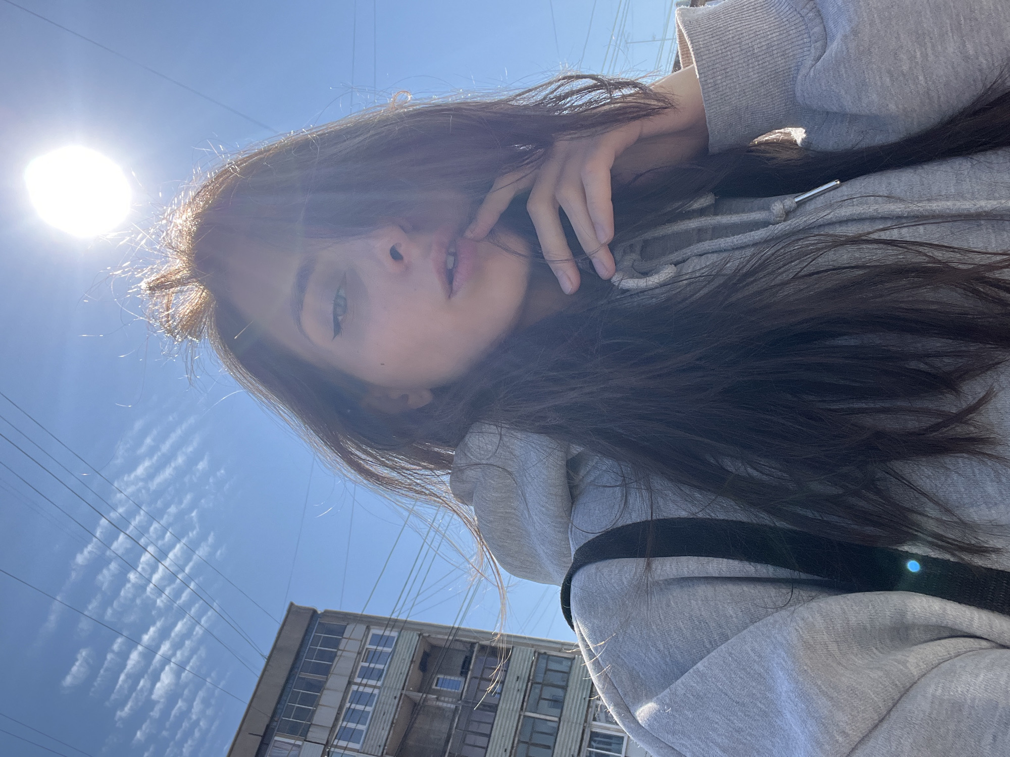 A young woman with dark hair is positioned in the foreground, partially obscuring the sun. She is wearing a grey hoodie with a black strap visible. Her hand is near her face. The background consists of a bright blue sky with wispy clouds and overhead power lines. A multi-story building with rows of windows is visible in the lower portion of the frame. It is possible this picture was taken in Krasnoyarsk, Russia, but it cannot be definitively determined from this picture.