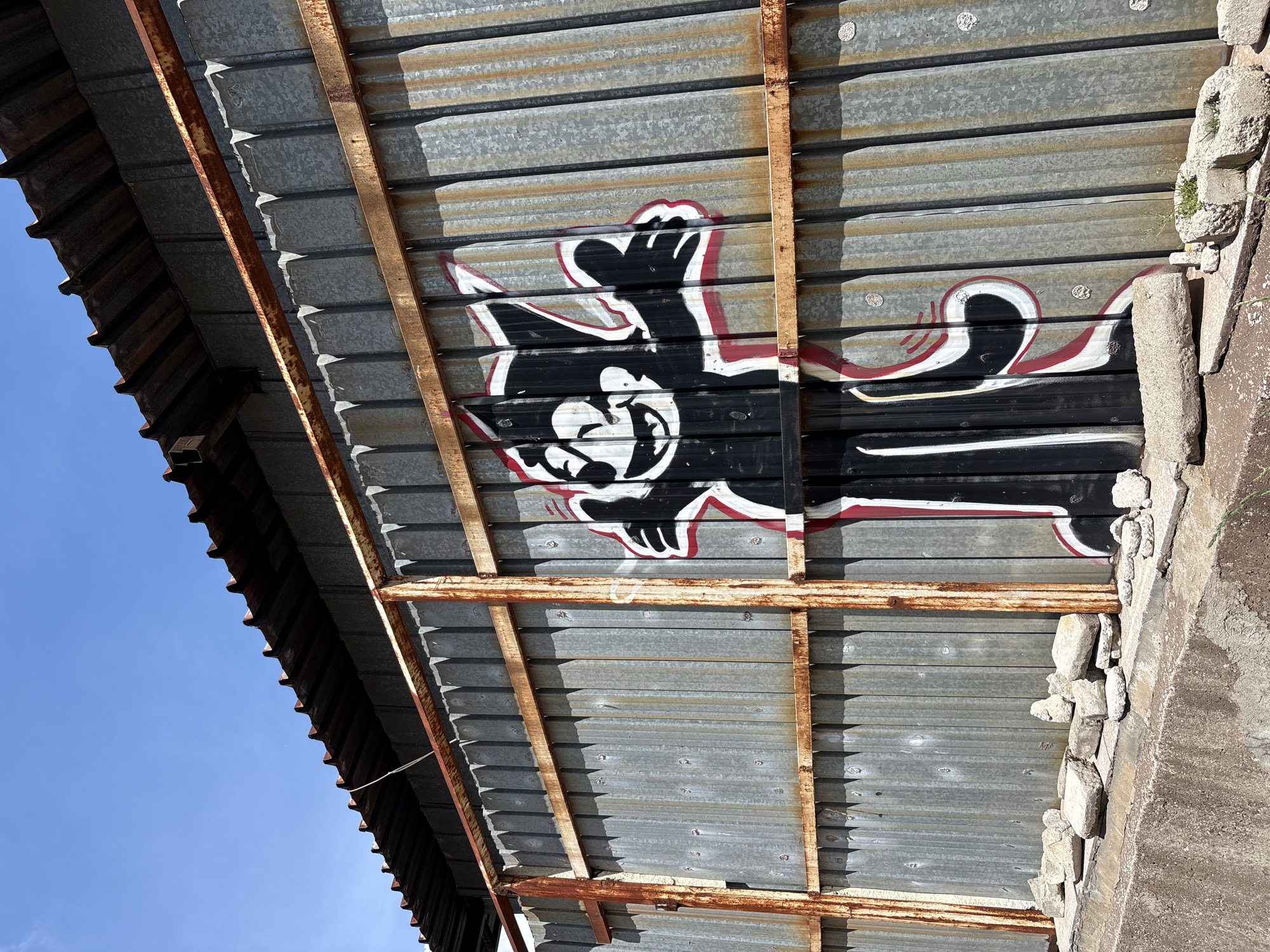 A low-angle shot depicts the underside of a corrugated metal roof supported by a rusty, metal frame. Graffiti of a black cartoon cat with white and red accents adorns the metal. The roof is affixed to a concrete structure with uneven stones. A clear blue sky is visible in the lower-left corner. The location is Sofia, Bulgaria.