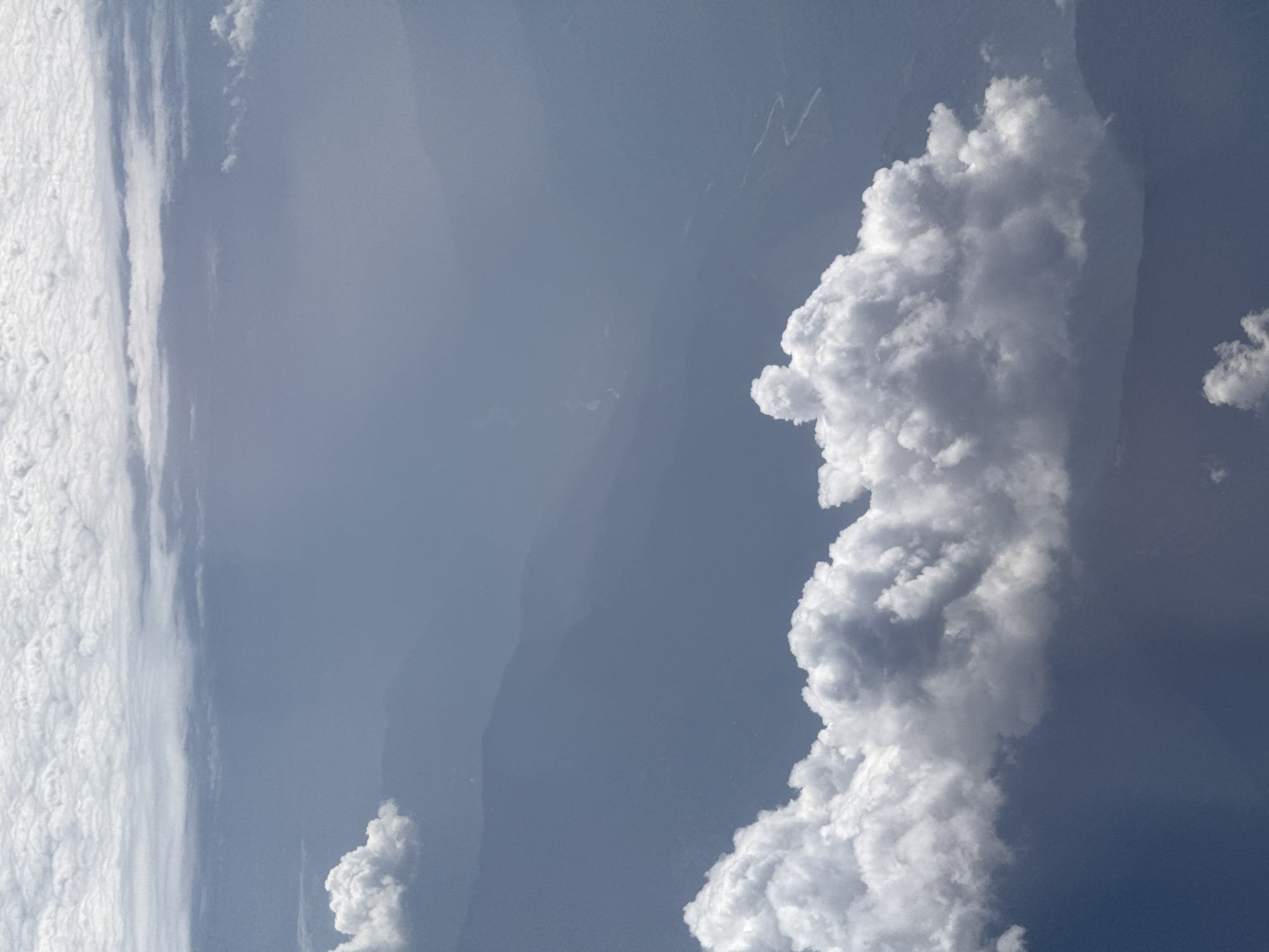 This is a photograph of clouds seen from above, likely from an airplane or satellite, given the perspective. The image captures a vast expanse of the sky, with a prominent, towering cumulus cloud formation dominating the right side of the frame. This cloud is bright white and exhibits a detailed, fluffy texture, suggesting it is a substantial cloud, possibly a cumulonimbus. To the left, a less defined layer of stratiform clouds stretches across the image, appearing as a thick, textured blanket.
The background is a hazy, muted blue sky, with hints of landforms faintly visible beneath the cloud cover, suggesting the photograph was taken at a high altitude. These landforms appear mountainous and rugged, with some areas appearing darker and others lighter, possibly indicating valleys or exposed rock faces. There are also thinner, wispy cloud streaks or contrails visible in the upper portion of the sky.
The lighting suggests it is daytime, with the sun illuminating the clouds from above, creating bright highlights and subtle shadows within the cloud structures. The overall mood is serene and awe-inspiring, highlighting the grandeur of atmospheric formations. The location context of Leh, India, is consistent with the mountainous terrain visible, and the clear, high-altitude skies often seen in that region. No text is visible in the image.