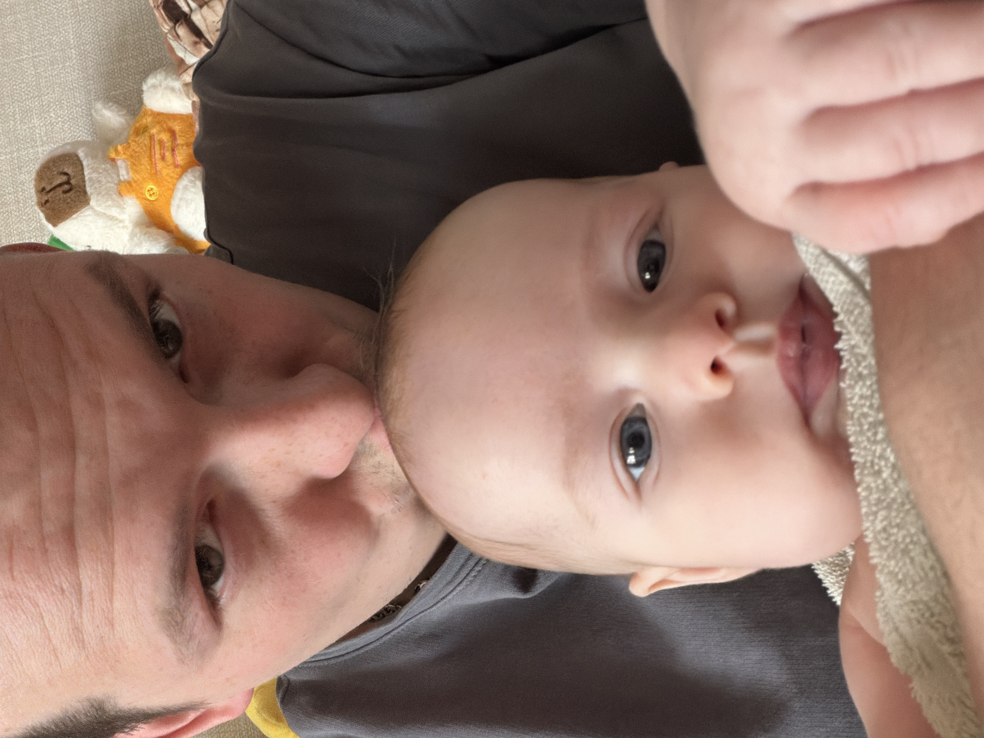 A selfie shot depicts a man and a baby. The man's face is visible on the left, with light skin, brown hair, and brown eyes. He is wearing a grey shirt. To the right, a baby with light skin, blue eyes, and a bald head is seen, with their tongue slightly protruding. The baby is partially obscured by a hand holding a grey towel. A stuffed animal is visible in the background, on the upper left. The location is unknown, and there is no evidence to suggest the city of Chaikovsky, Russia, is related.