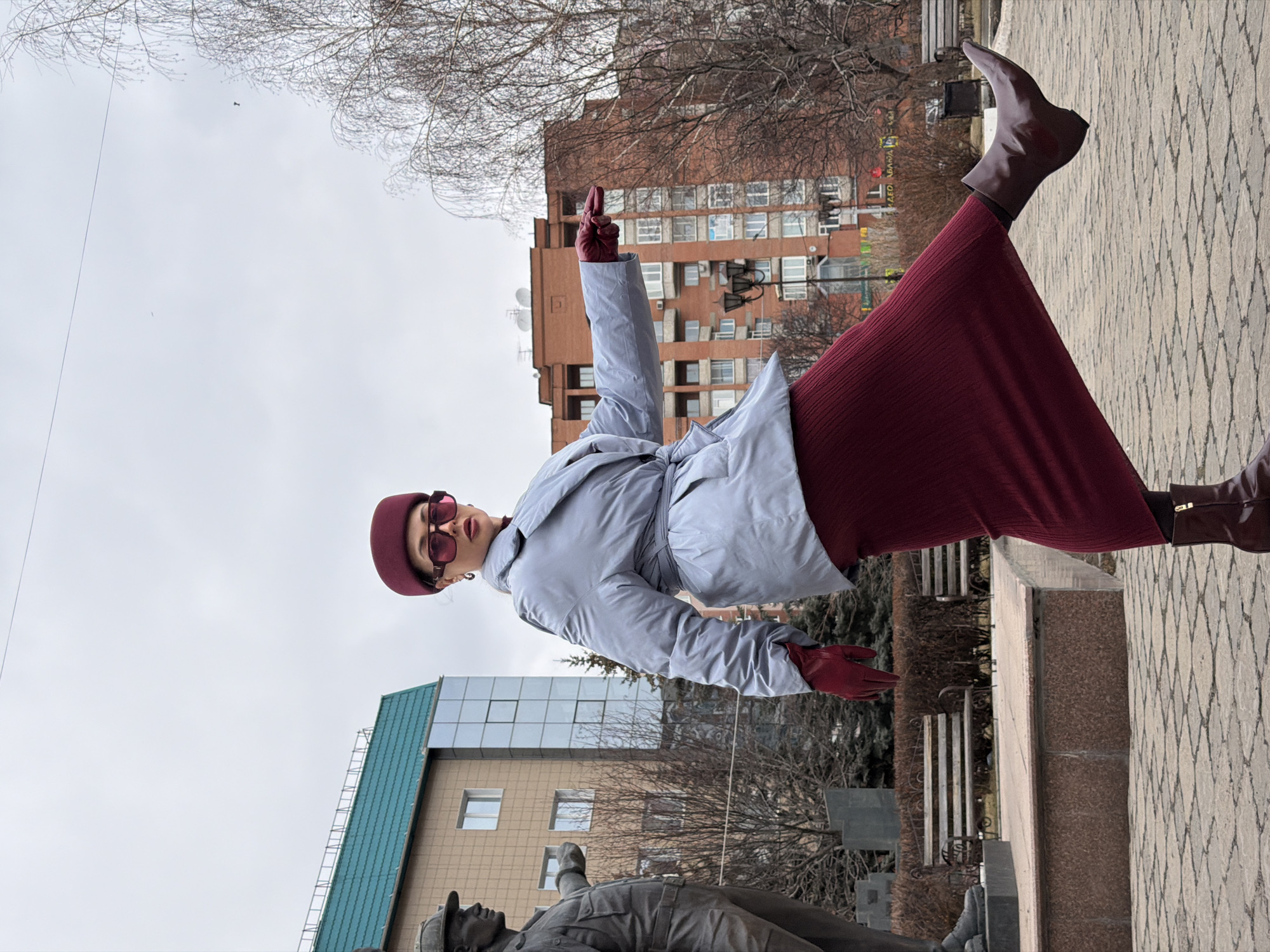 A full-length shot depicts a woman posing in front of a monument in Krasnoyarsk, Russia. She wears a burgundy beret, matching gloves, dark-framed sunglasses, a light grey trench coat with a tie around the waist, a burgundy knit dress, and brown leather ankle boots. Her right arm is raised skyward with her index finger extended. The monument is in the foreground, showing a partially obscured bronze figure of a man in uniform. Behind the woman, the cityscape reveals residential buildings under an overcast sky. The ground is covered with a patterned pavement. The scene includes architectural details such as windows, balconies, and building facades, with sparse trees and branches visible in the background. A statue is also in view.