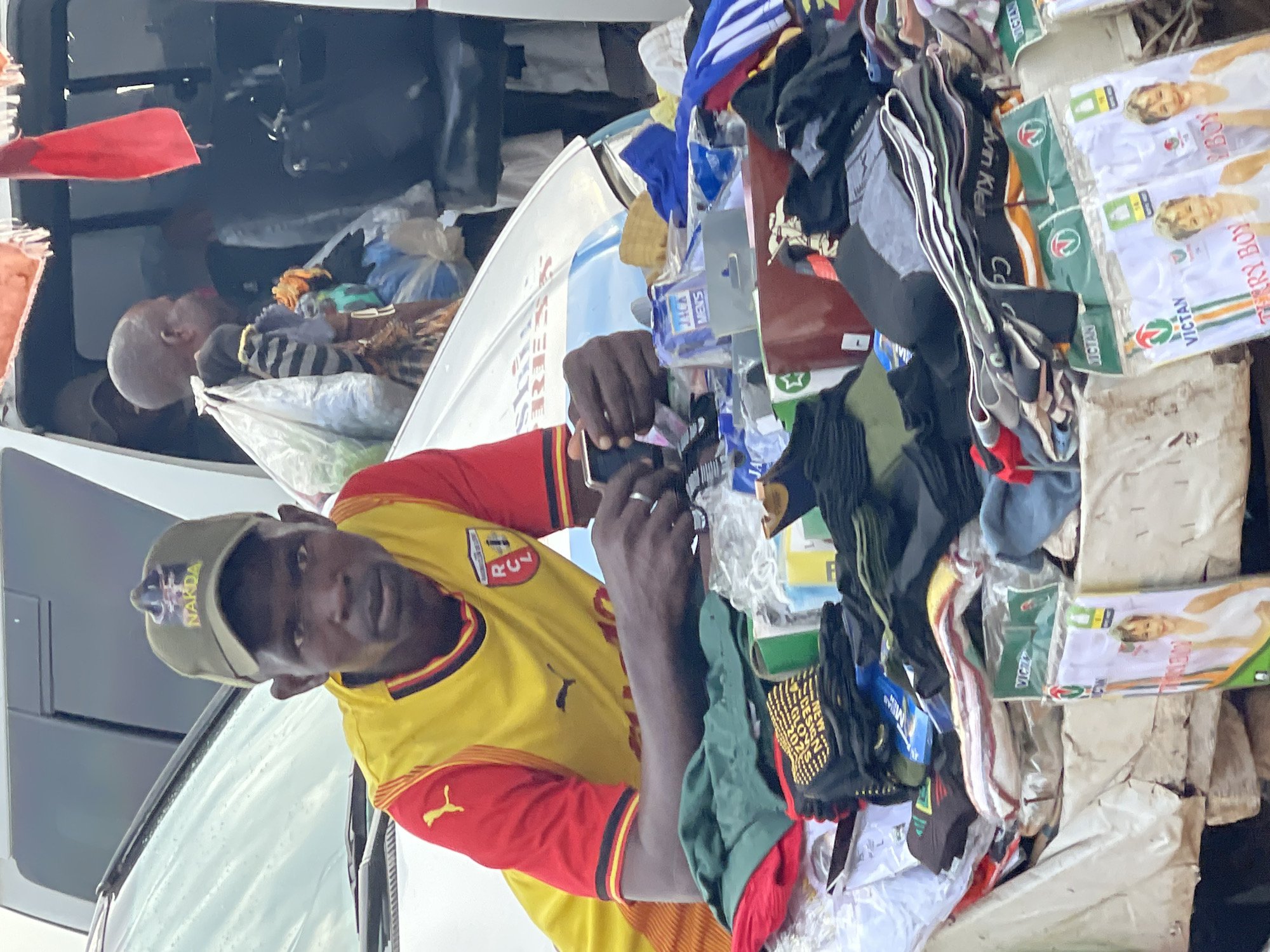 A Black man wearing a yellow and red short-sleeved jersey with a green baseball cap is shown in the foreground, sitting on the hood of a white vehicle, facing the camera. He has a neutral expression. His hands are visible, with the right hand holding a dark-colored smartphone, and the left hand resting on a pile of merchandise. The merchandise consists of folded clothing items, including socks and undergarments, some of which are in clear plastic packaging with text such as "VICTON," "CALVIN KLEIN," and "NEW DESIGN GLOVES 2023." The vehicle's hood, which is white and has some faint blue lettering that is difficult to decipher, is covered with this merchandise. In the background, another Black person is visible partially through the open door of the vehicle, carrying several bags. The scene appears to be outdoors, possibly a market stall or roadside vendor setup. The overall lighting suggests it is daytime, with some overcast conditions. The presence of the specific merchandise packaging, such as "VICTON" which is a known brand in Nigeria, and the general atmosphere suggest the scene is likely located in an urban setting within Nigeria. Given the provided information, it is reasonable to infer the location is Abuja, Nigeria.