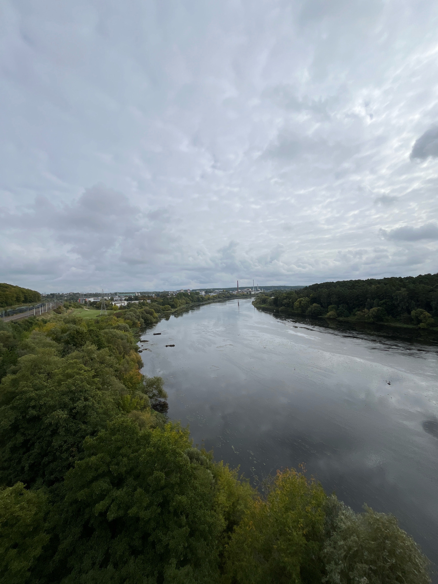 This is an elevated, wide-angle view of the Nemunas River in Kaunas, Lithuania, on a cloudy day. The river takes up the majority of the frame, its dark, reflective surface showing ripples and patches of debris or algae. On the left bank, a dense area of green trees lines the river, with a railway line and industrial buildings visible further inland. On the right bank, a thick forest extends along the river's edge, with more distant buildings and structures appearing on the horizon. The sky is overcast with a textured pattern of gray clouds, suggesting a mild, possibly cool, climate. There are no people visible in the image. A few dark, rectangular objects, possibly buoys or debris, are scattered in the water near the left bank.