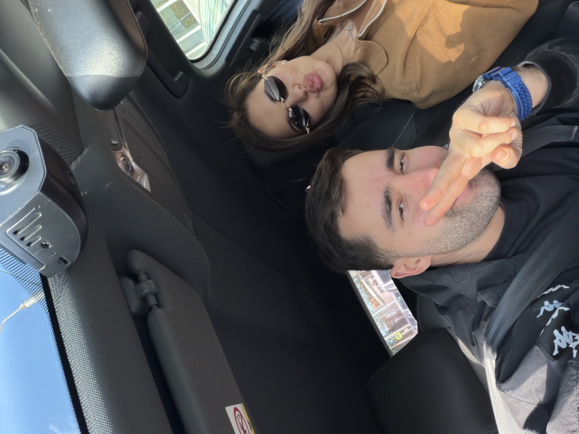 A man and a woman are in the front seats of a car. The man is wearing a blue watch on his left wrist, a black jacket with Kappa logos, and has a trimmed beard. He is holding up two fingers in a V-shape. The woman, who is wearing sunglasses, is lying back in her seat and pouting. She is wearing a brown jacket and has long brown hair. The scene is inside the vehicle.