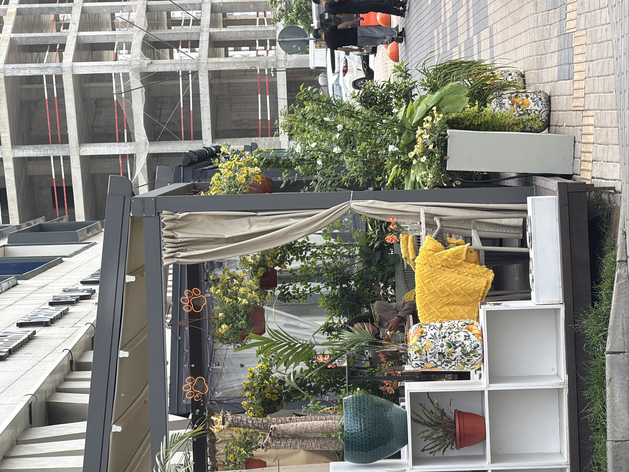 The image captures an outdoor seating area of a establishment in an urban environment, likely in the city of Unknown, Unknown. In the foreground, a low white shelving unit displays a potted fern on the left and a decorative vase filled with a citrus-themed pattern on the right. Next to the shelving unit, a bright yellow textured blanket is draped over a chair. Behind the seating area, lush green plants and hanging baskets of yellow flowers create a vibrant backdrop. A beige curtain hangs from a canopy overhead. In the mid-ground, a person is seated at a table, looking at their phone. Further back, several cars are parked on a paved walkway, and two men are walking away from the camera. In the background, a large, unfinished concrete building dominates the skyline, indicating ongoing construction. The ground is a patterned brick walkway.