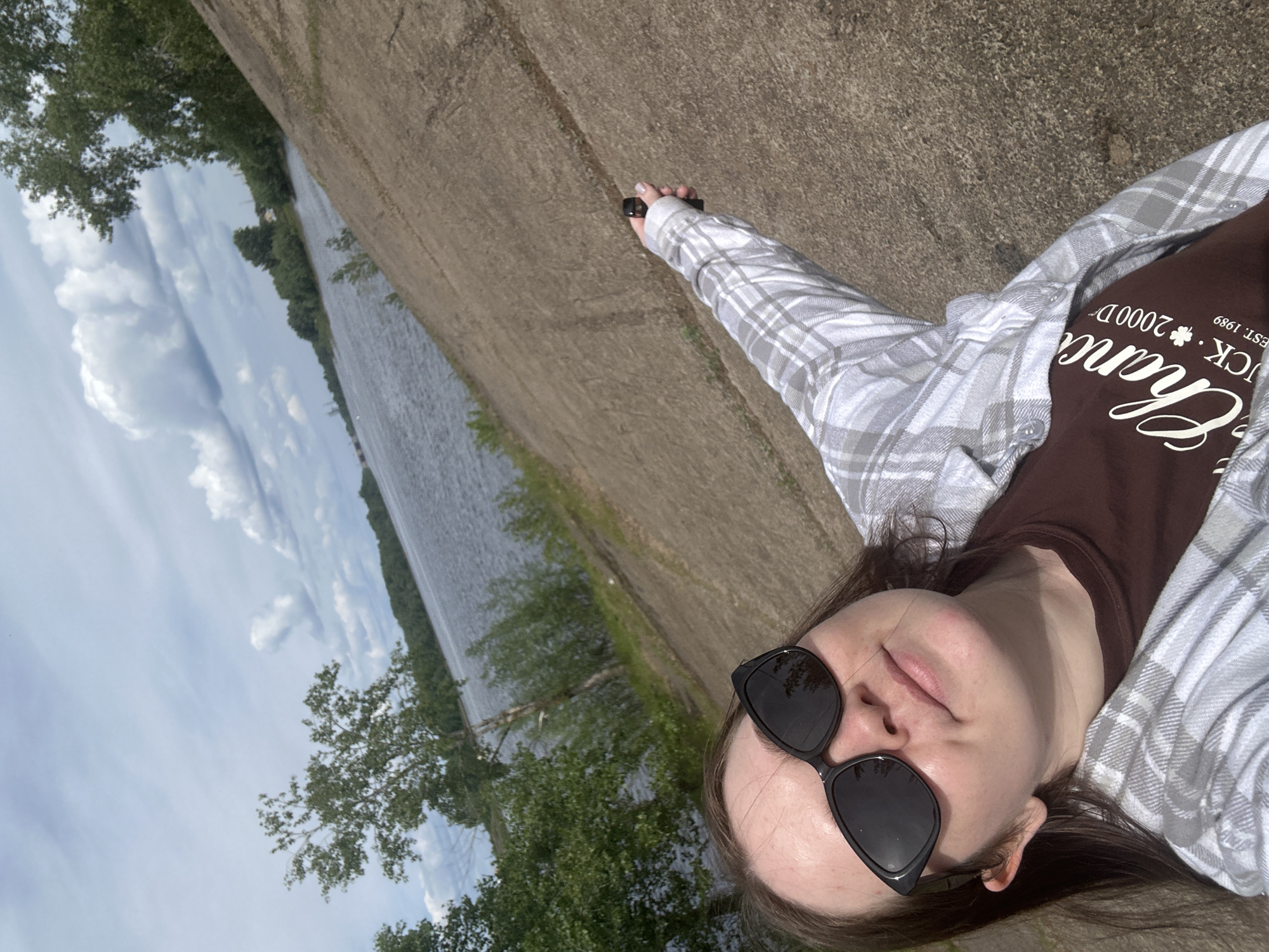 The image, originally oriented on its side, depicts a person lying on a concrete or paved embankment next to a wide body of water, likely a river or a large canal, in Chaikovsky, Russia.
In the foreground, a young female is seen, facing the camera with her head slightly tilted. She wears black, rectangular sunglasses that obscure her eyes. Her dark hair frames her face. She is dressed in a light-colored, grey and white plaid flannel shirt, worn unbuttoned over a brown short-sleeved t-shirt. On the brown t-shirt, inverted text is visible, which when rotated reads "Champ" or "Chance" followed by "CK", "EST. 1989" and "2000 D". Her left arm is extended, and she holds a small, dark, rectangular object in her hand, possibly a vape pen or a remote control.
The background shows the expansive water body, which has small ripples on its surface. The far bank is lined with lush green trees. Above, the sky is bright blue with scattered white, fluffy cumulus clouds, indicating a clear and sunny daytime. The overall scene suggests a relaxed outdoor setting, possibly during warm weather, with the person enjoying the view while taking a selfie or resting.
