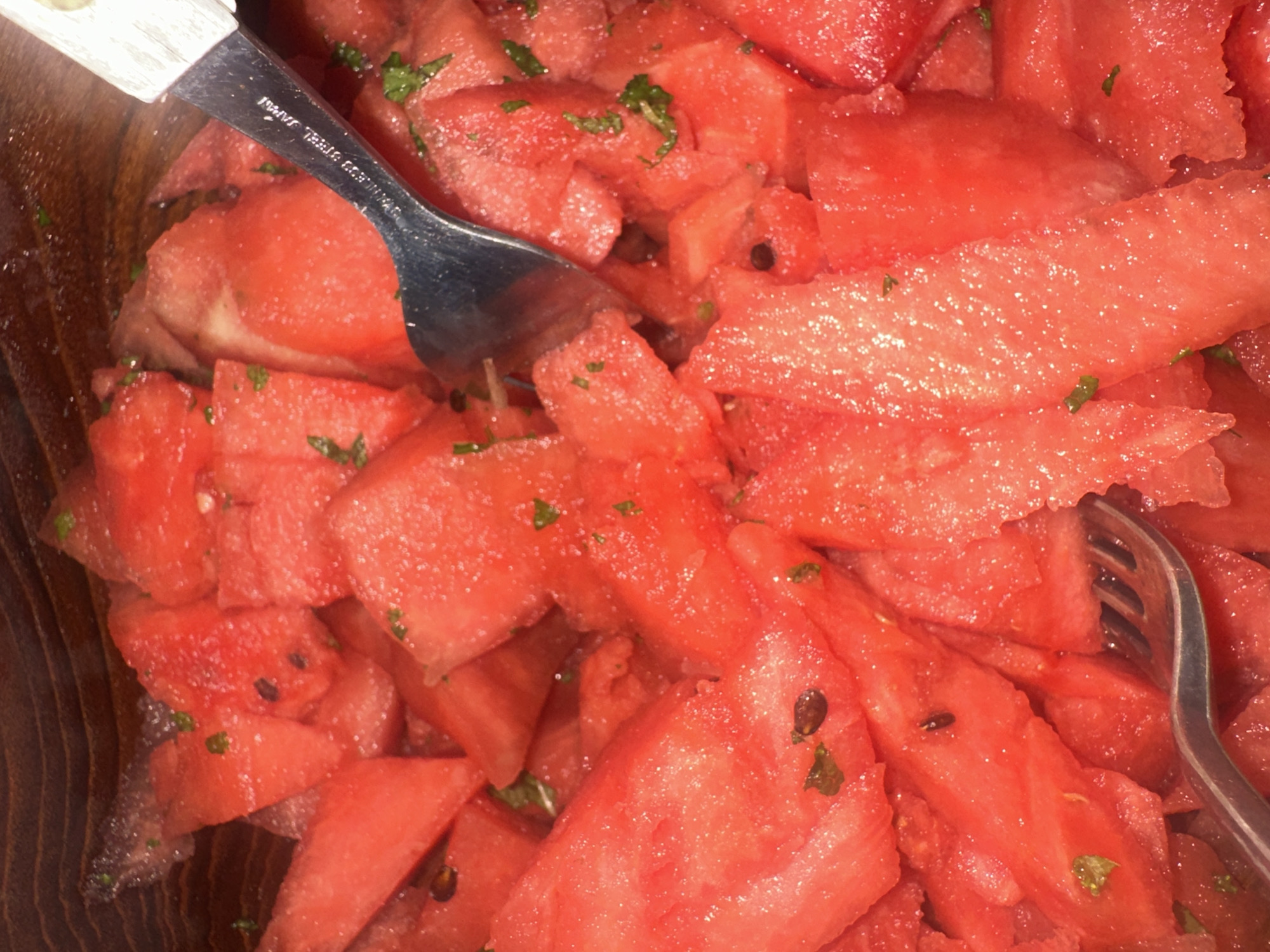 This close-up image presents a vibrant bowl of cut watermelon, appearing fresh and juicy. The watermelon pieces are predominantly bright red, irregularly cut into chunks and triangles, and some black seeds are visible. Scattered throughout the fruit are small green specks, likely chopped mint or basil, suggesting a refreshing fruit salad.
Two serving utensils are visible within the bowl. A metal fork with a white handle is partially submerged in the watermelon at the top left, with the text "STAINLESS STEEL JAPAN" clearly engraved on its shaft. Another entirely metal fork is visible on the right side of the frame, also partially resting among the fruit. The presence of these forks indicates that the watermelon is either being served or consumed.
The fruit is contained within a dark wooden bowl or serving dish, whose textured grain is visible along the left and lower edges of the image. The lighting appears to be indoor, possibly artificial, reflecting off the moist surfaces of the watermelon. There are no people visible, and no specific location cues for London, United Kingdom, are discernible from the image content itself, nor can the time of day or weather be determined.