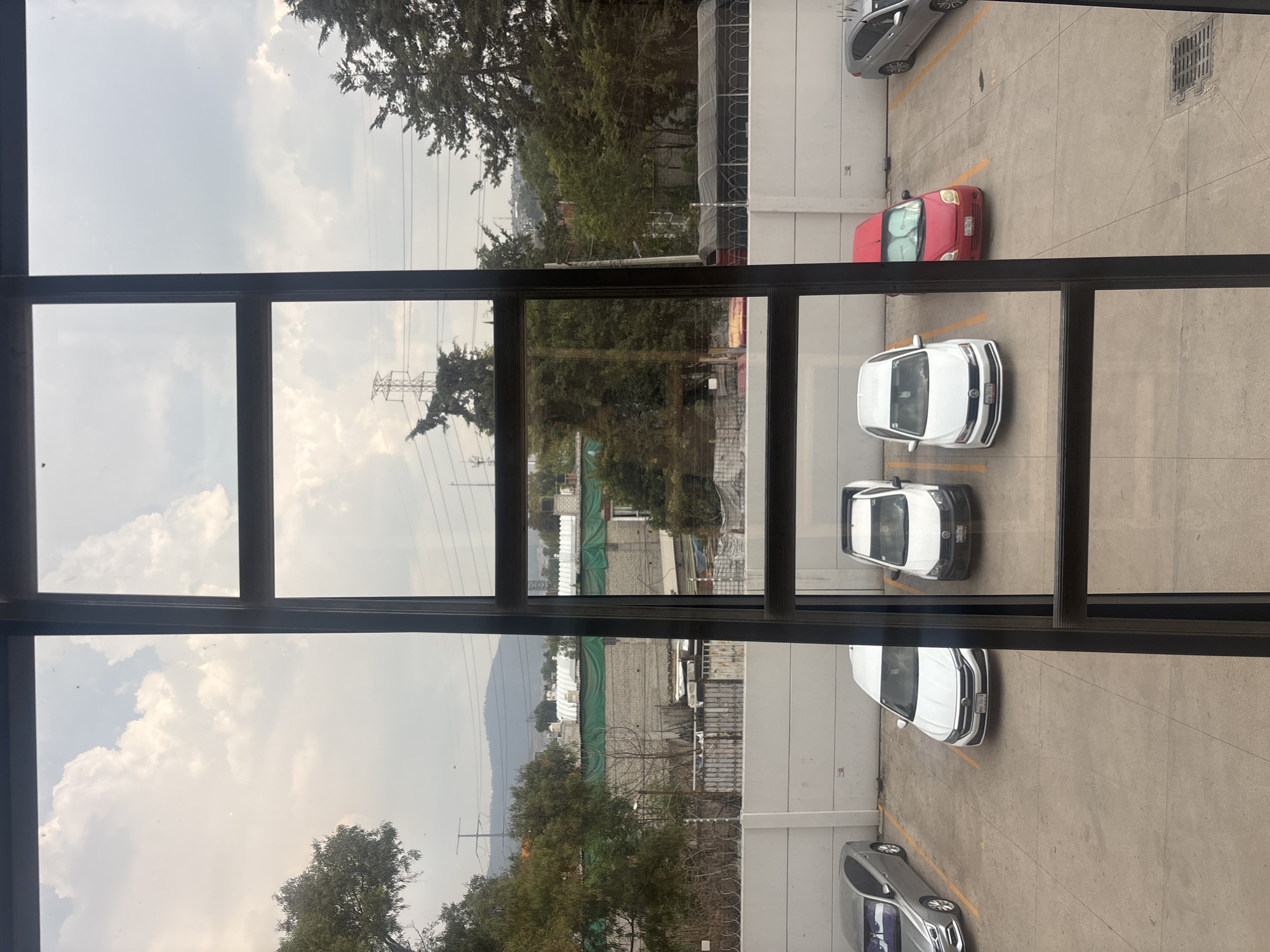 The image is taken from an elevated position looking through a multi-paned window in Buenavista, Mexico. Outside, there is a parking lot. It contains several cars: two white compact sedans, a gray crossover SUV, and a red compact car. There are trees and a building visible in the background, beyond the parking lot. The sky is overcast. High-tension power lines are also visible. There are no people visible.