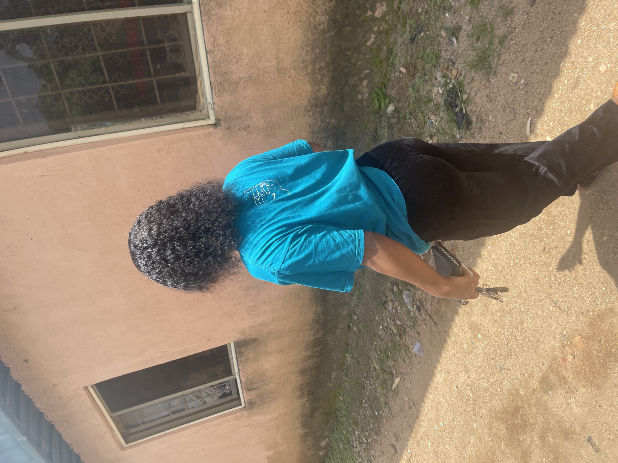 A person walks away from the camera in a dusty outdoor environment. The person is wearing a bright blue t-shirt with a white graphic design on the back and black pants. They are holding a black wallet and a set of keys in their right hand. The background consists of a light pink building with two windows. The ground is covered in dirt and small stones. This scene appears to be located in a residential area within the city of Jos, Nigeria, evidenced by the style of the building, the dusty ground, and the general ambiance typical of many neighborhoods in Jos.