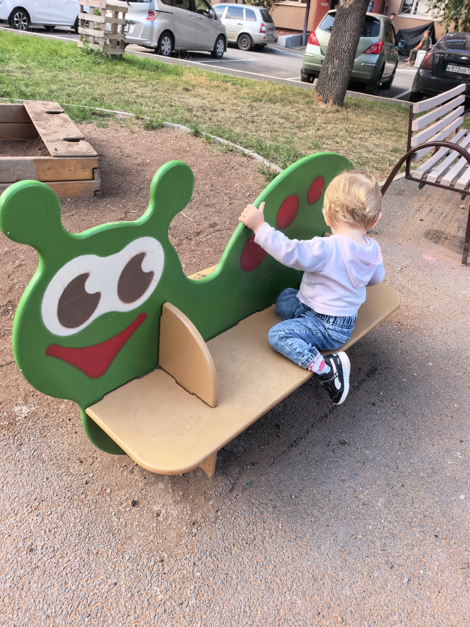 In Sterlitamak, Russia, a young child, viewed from the back, sits on a green caterpillar-shaped playground ride. The child wears a light pink hoodie and blue jeans, with black sneakers on their feet. The playground equipment is made of brightly colored plastic and metal, with a beige seat. The background shows a gravelly ground, grass, trees, parked cars, and a residential building.