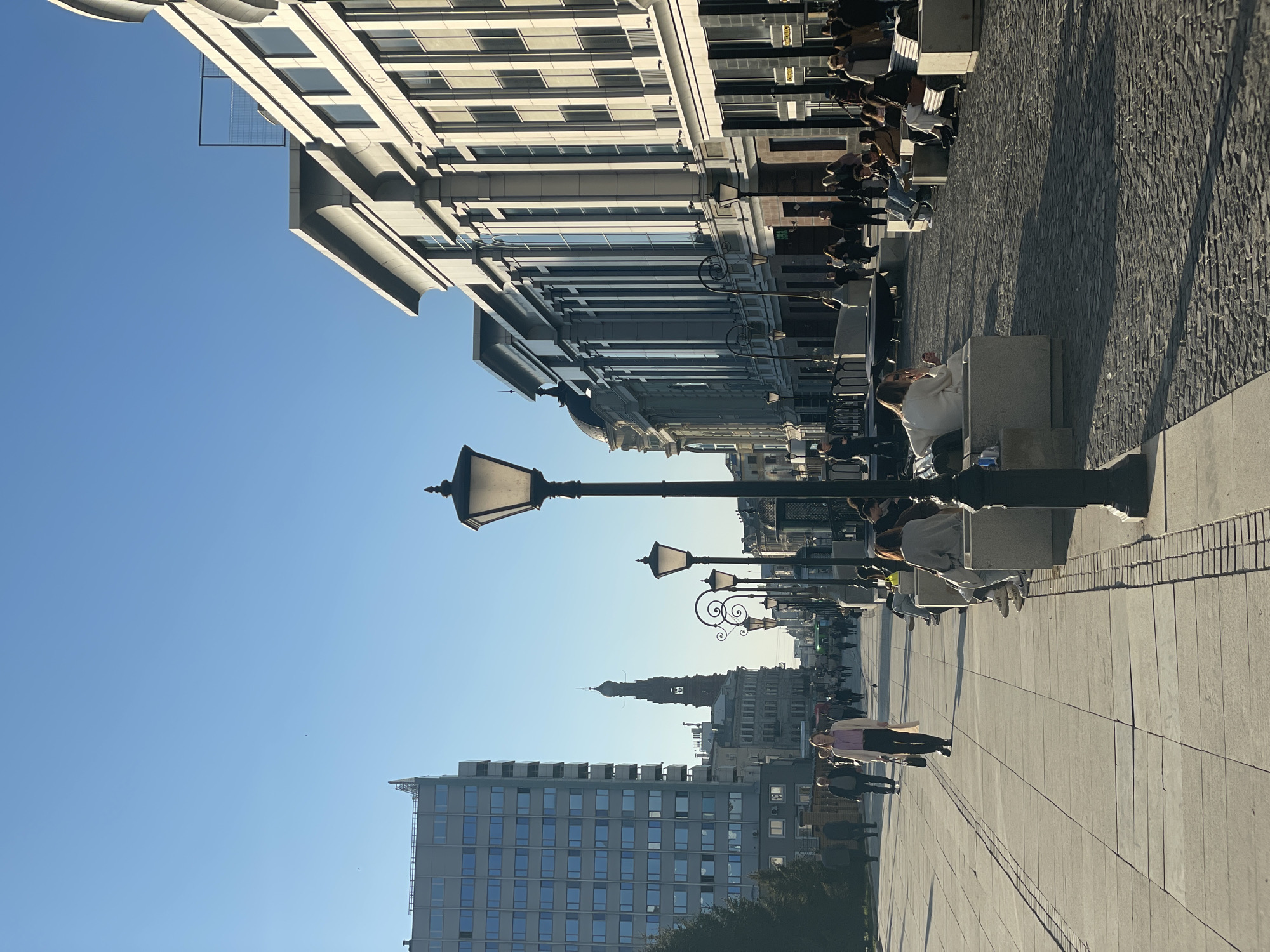 The image depicts a pedestrian street in Kazan, Russia. Along the street are benches, lampposts, and buildings of varied architectural styles, ranging from modern to traditional. People are seen walking and sitting on benches. The sky is clear and blue. The scene suggests a sunny day in the city. The street is paved with large, light-colored stones, and the adjacent pedestrian area is paved with smaller, darker stones.