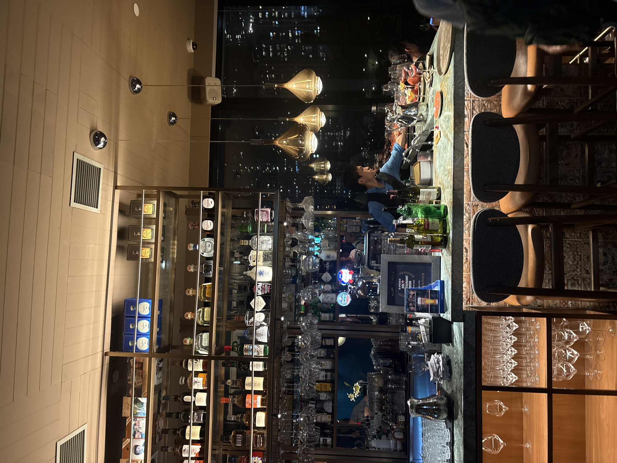 The image shows the interior of a bar, possibly in Hong Kong, China, given the high-rise cityscape visible through the windows. The scene is primarily focused on the bar area, where a bartender is seen working. He is wearing a blue shirt and appears to be preparing drinks. The bar is well-stocked with a variety of alcoholic beverages displayed on shelves behind the counter. Numerous bottles of liquor and various glassware are arranged neatly. Above the bar, there are pendant lights hanging down. Patrons are seated at the bar on high stools. The stools have dark seats and wooden frames. The bar counter is marble-like, and there are some appetizers and drinks on it. A display case with glasses is located below the bar area. Overall, the scene depicts a typical evening ambiance in a bar, presumably located in a high-rise building in Hong Kong.