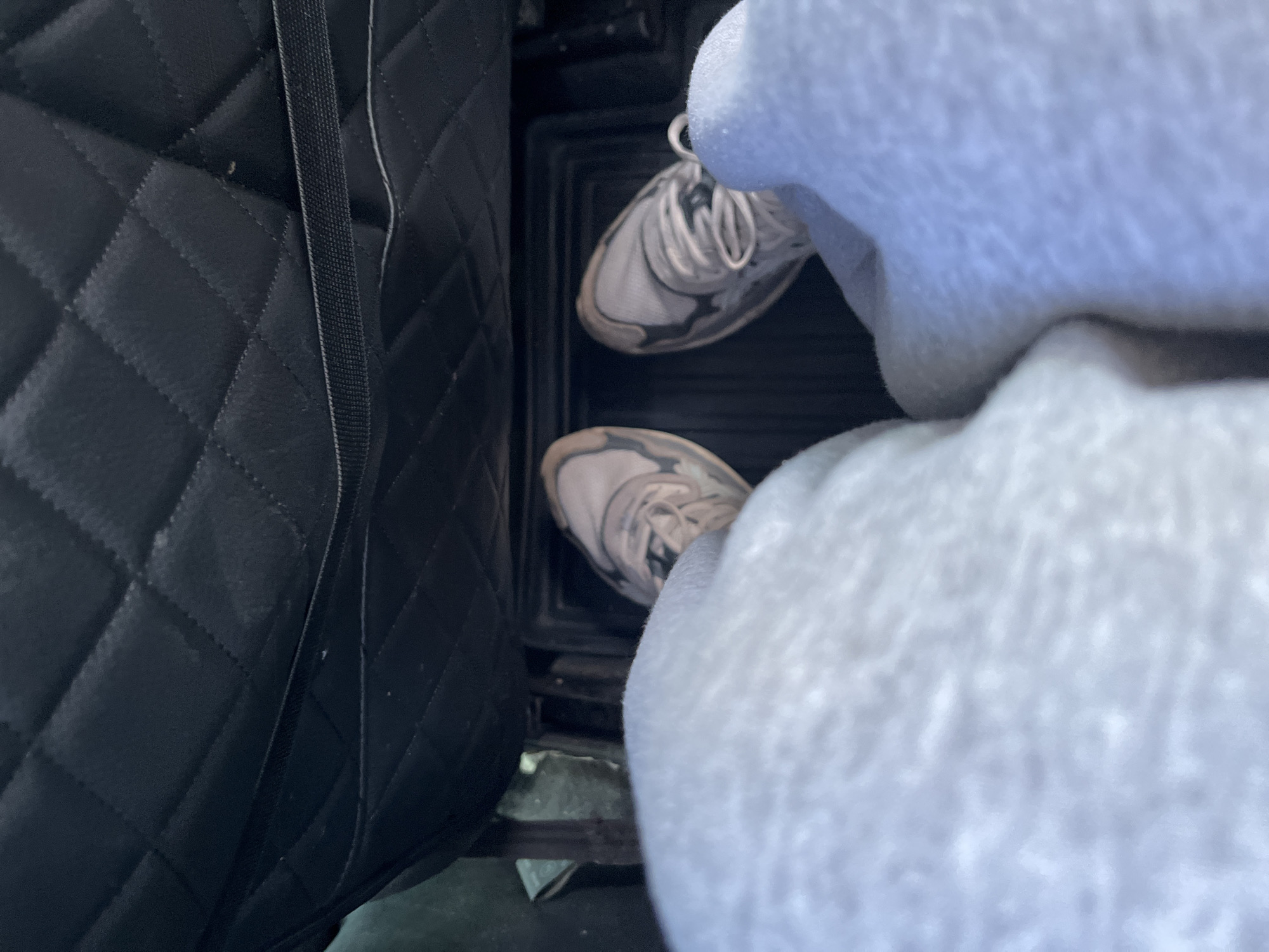 The image shows a person's legs and feet inside a vehicle. The person is wearing light gray sweatpants and white sneakers with dark gray accents. The feet are positioned on a black floor mat. Behind the feet, the interior of the vehicle is visible, including a quilted black seat or panel. The scene appears to be indoors, possibly in a parked vehicle. There is no discernible information about the city of Марьевка, Russia, within the image itself.