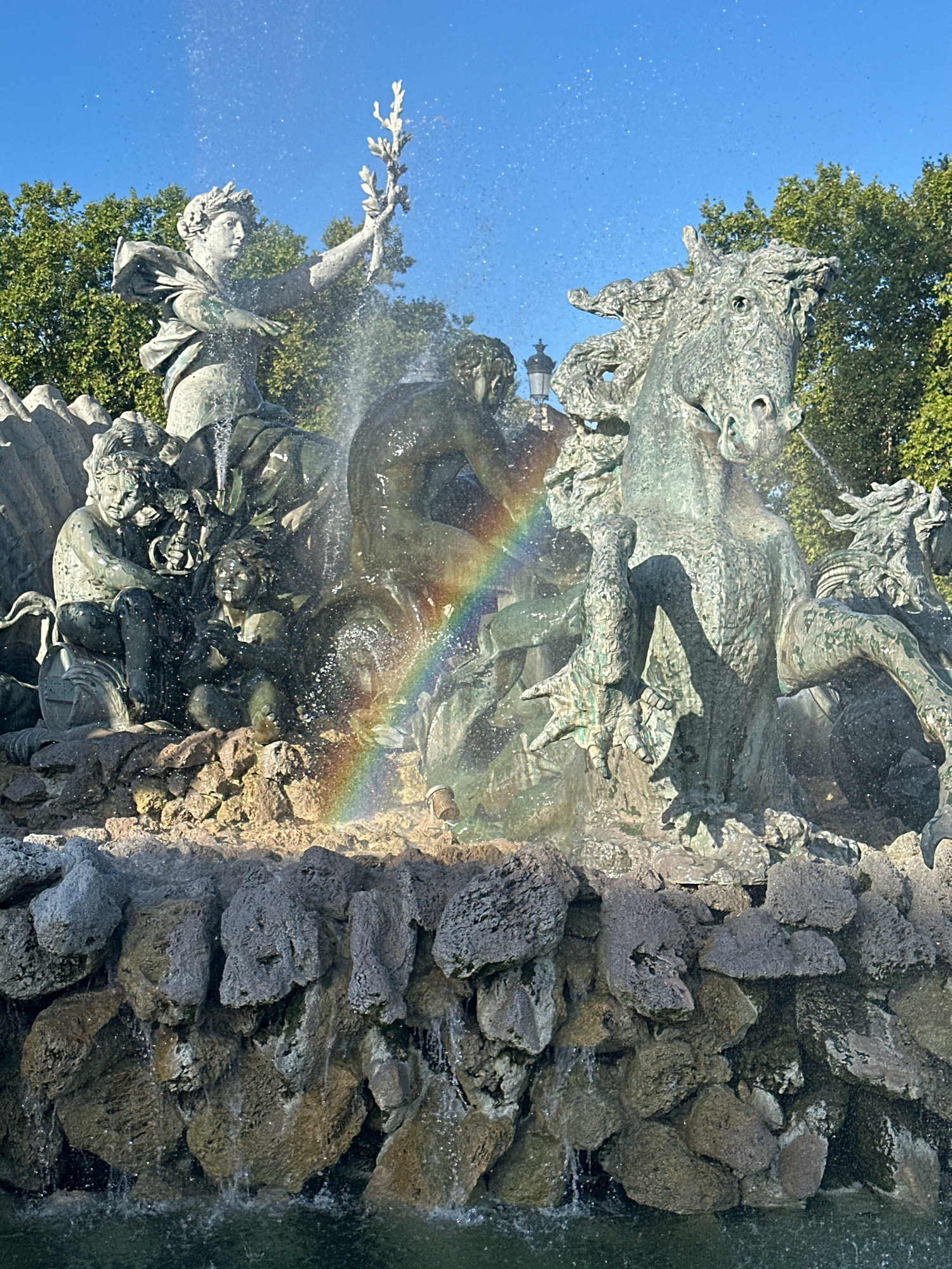 This is a close-up, eye-level shot of the "Fontaine des Girondins" in Bordeaux, France. The fountain, made of bronze and stone, is the central subject. The upper part of the fountain features a female allegorical figure wearing a laurel wreath, holding a staff with a leafy branch. She is surrounded by water spray, through which a rainbow is visible. To her right, the head and mane of a horse are depicted, with water erupting from its mouth and mane. Below, a group of figures, possibly children or putti, are incorporated into the rocky base of the fountain. The fountain is set against a clear blue sky and green trees in the background. The water cascades over a textured, rocky base into a pool at the bottom, which is partially visible. The overall lighting suggests it is daytime, with sunlight illuminating the scene and creating highlights on the water and bronze.