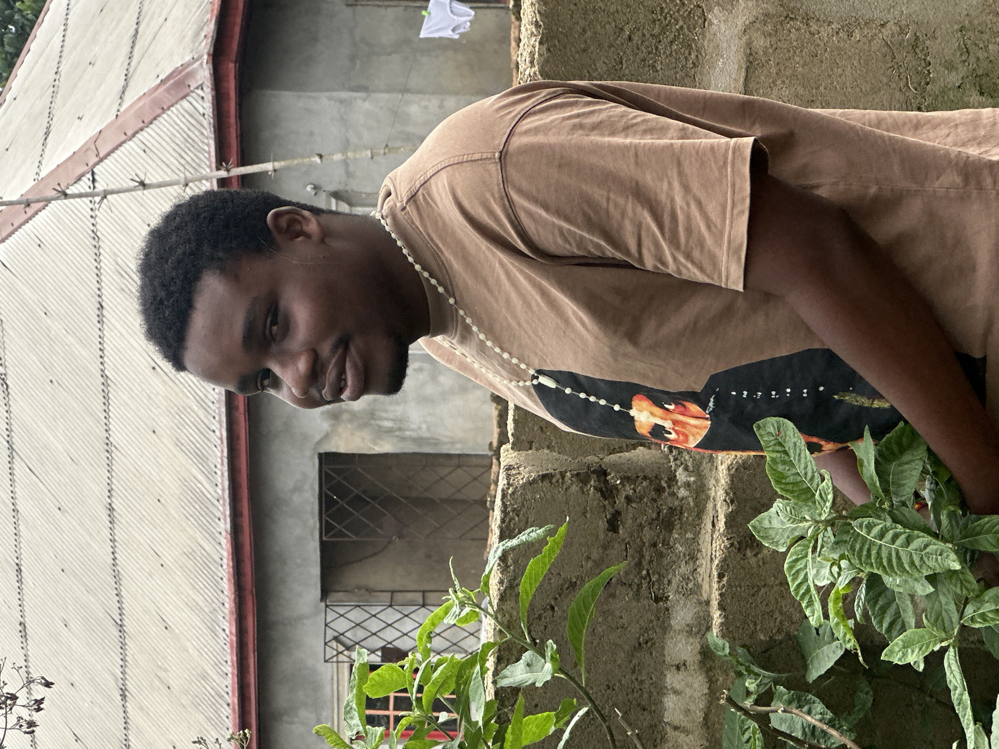 A young man stands outdoors in Ikot Ekpene, Nigeria. He is wearing a brown t-shirt with a graphic design and a beaded necklace. The setting includes a building with a corrugated metal roof, a barred window, and a rough concrete wall. Plants are growing at the base of the wall. The man is looking down at the plants. A white cloth is hanging on a line.