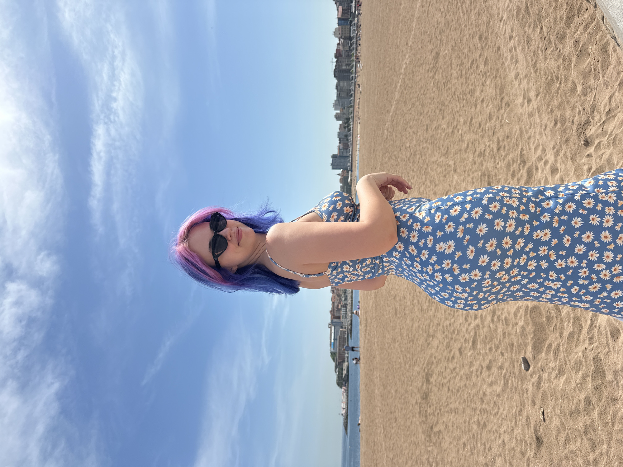A portrait-style photograph captures a young woman standing on the beach of Gijón/Xixón, Spain. She is positioned to the left, sporting sunglasses and a floral blue dress. Her hair is dyed with purple and blue hues. The backdrop features a clear blue sky dotted with sparse clouds to the left, while the expansive sandy beach stretches out to the right, bordering a cityscape in the distance. The shoreline, with the sea visible, lies between the beach and the city buildings.
