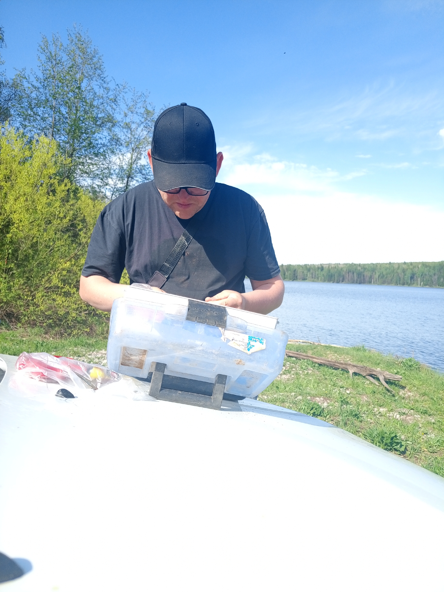 A man wearing a black T-shirt, black baseball cap, and glasses is standing in front of a clear plastic tackle box, which is sitting on a white surface (possibly a boat or vehicle). He is looking down at the box and appears to be organizing or inspecting the contents. To the left of the tackle box are some items in plastic bags and a small, dark object. The background consists of a body of water (possibly a lake) and a treeline on the opposite shore. To the left of the man is a patch of green vegetation, including trees. The sky is blue with some scattered clouds. The location is Уральское, Russia, based on available information.