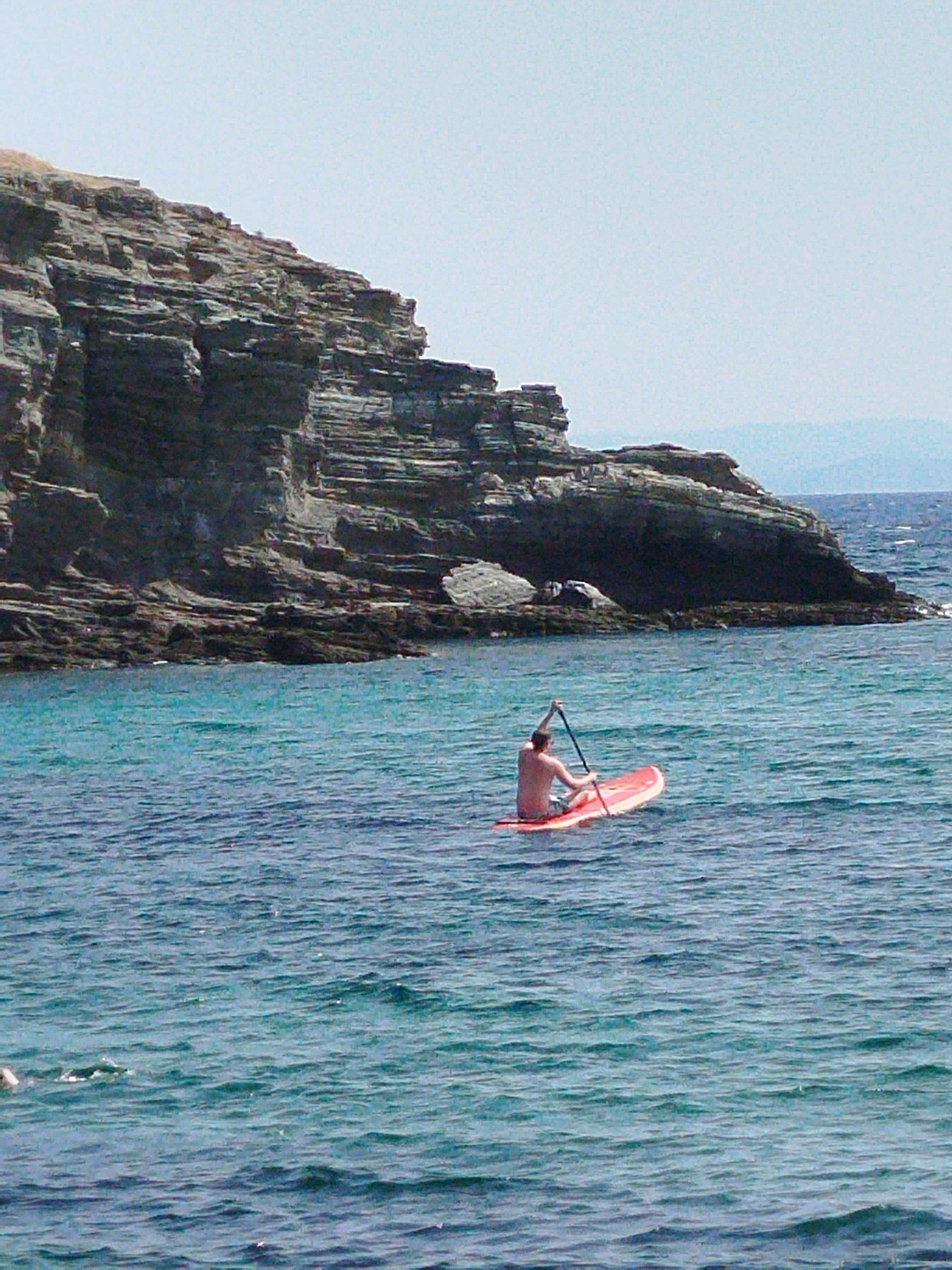 A person is paddleboarding in the sea near a rocky cliff. The person is seated on a red paddleboard and is holding a paddle. The sea is a vibrant blue-green color and is slightly choppy. The rocky cliff is a dark gray color and has a layered appearance. The sky is a pale blue and is clear. The image appears to be taken on a sunny day. The location is the coast of Sithonia Municipal Unit, Greece.