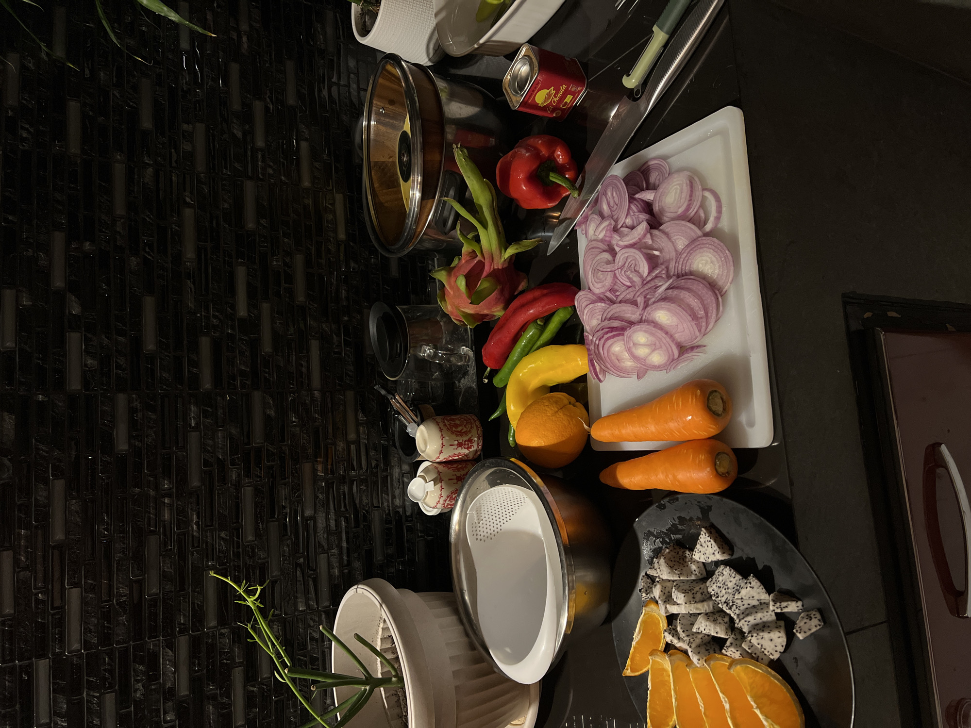 The image shows a kitchen counter with various fruits and vegetables prepared for cooking. There are sliced red onions on a white cutting board, along with whole carrots, a yellow bell pepper, and what appear to be chili peppers. Beside the cutting board, there is a black plate containing diced dragon fruit and sliced oranges. A metal bowl, possibly a strainer, sits near the plate. Other items include a can of La Chinata paprika, various kitchen utensils, and a potted plant. The counter has a dark, patterned surface. The location appears to be a domestic kitchen.