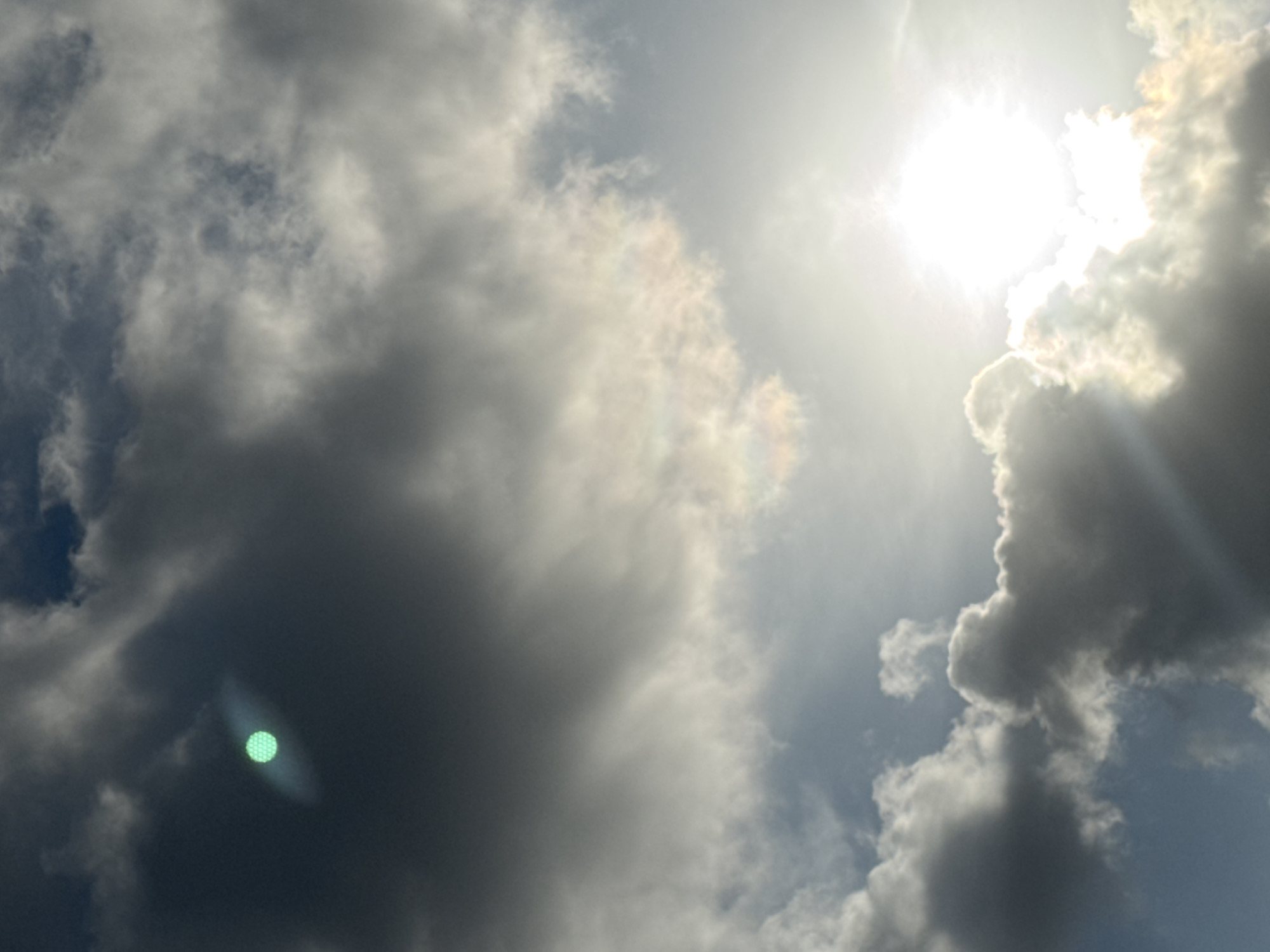 This is a photograph of the sky, featuring a dramatic interplay of dark and light clouds. In the upper left quadrant, a distinct circular object with a greenish hue is visible, surrounded by a diffused light effect. The sun is prominent in the lower right, its intense brightness obscured by a thick layer of clouds, casting light rays through the gaps. The overall scene suggests a potentially stormy or overcast sky with a localized atmospheric phenomenon. The image does not contain any discernible elements that would indicate the city of Karon, Thailand, or any specific urban or environmental context beyond a sky view.