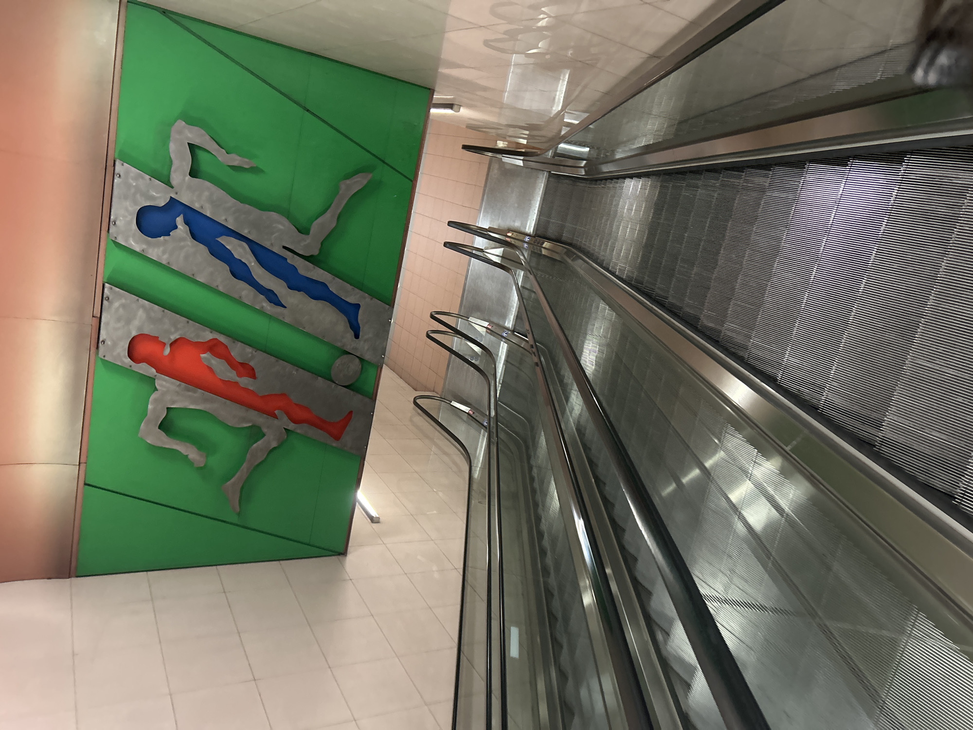 An eye-level interior shot shows a modern-looking structure with an escalator and wall art, probably taken in a public space such as a train station. The escalator takes up most of the right side of the image, starting from the upper right corner and descending diagonally towards the lower right. The escalator is metallic grey with a ribbed, moving platform. On the left side of the image, there is a bright green wall with two stylized metal cut-outs of a human body. One cut-out is filled with blue and the other is filled with red. The background wall is tiled with light-colored, shiny tiles. Based on the art style, this is likely the Konstantin Velichkov Metro Station in Sofia, Bulgaria.