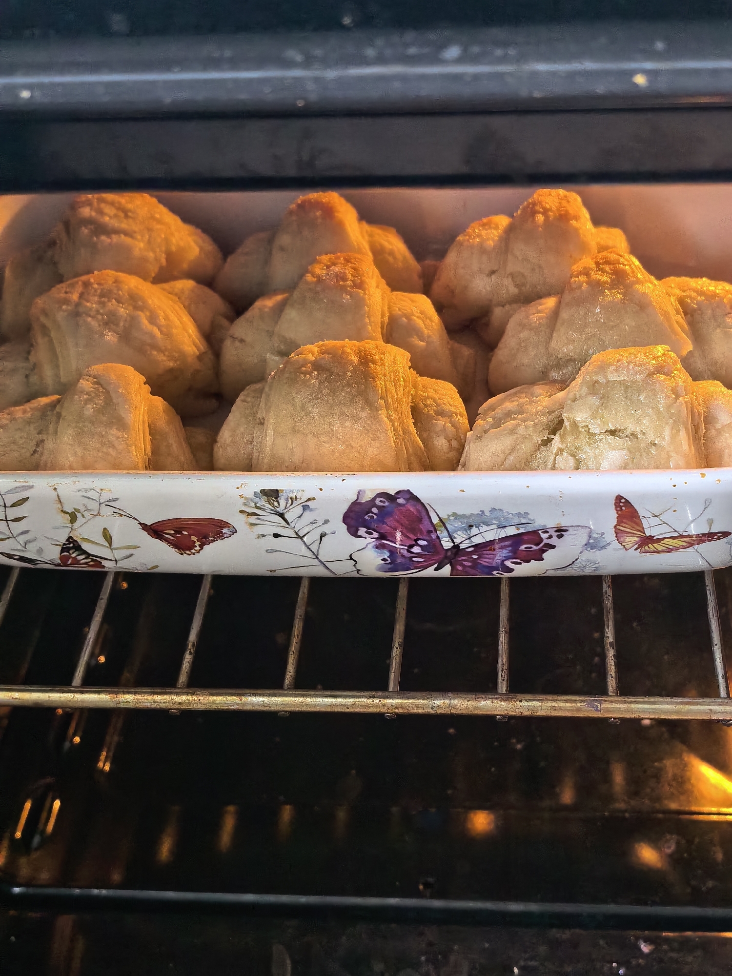 A baking dish filled with baked goods sits on a rack inside an oven. The dish is white with butterfly designs. The baked goods are crescent-shaped and appear to be golden brown. The inside of the oven is dark, with the heating element visible at the bottom. There are no people in the image. The photo does not provide any indication of location in Sterlitamak, Russia.