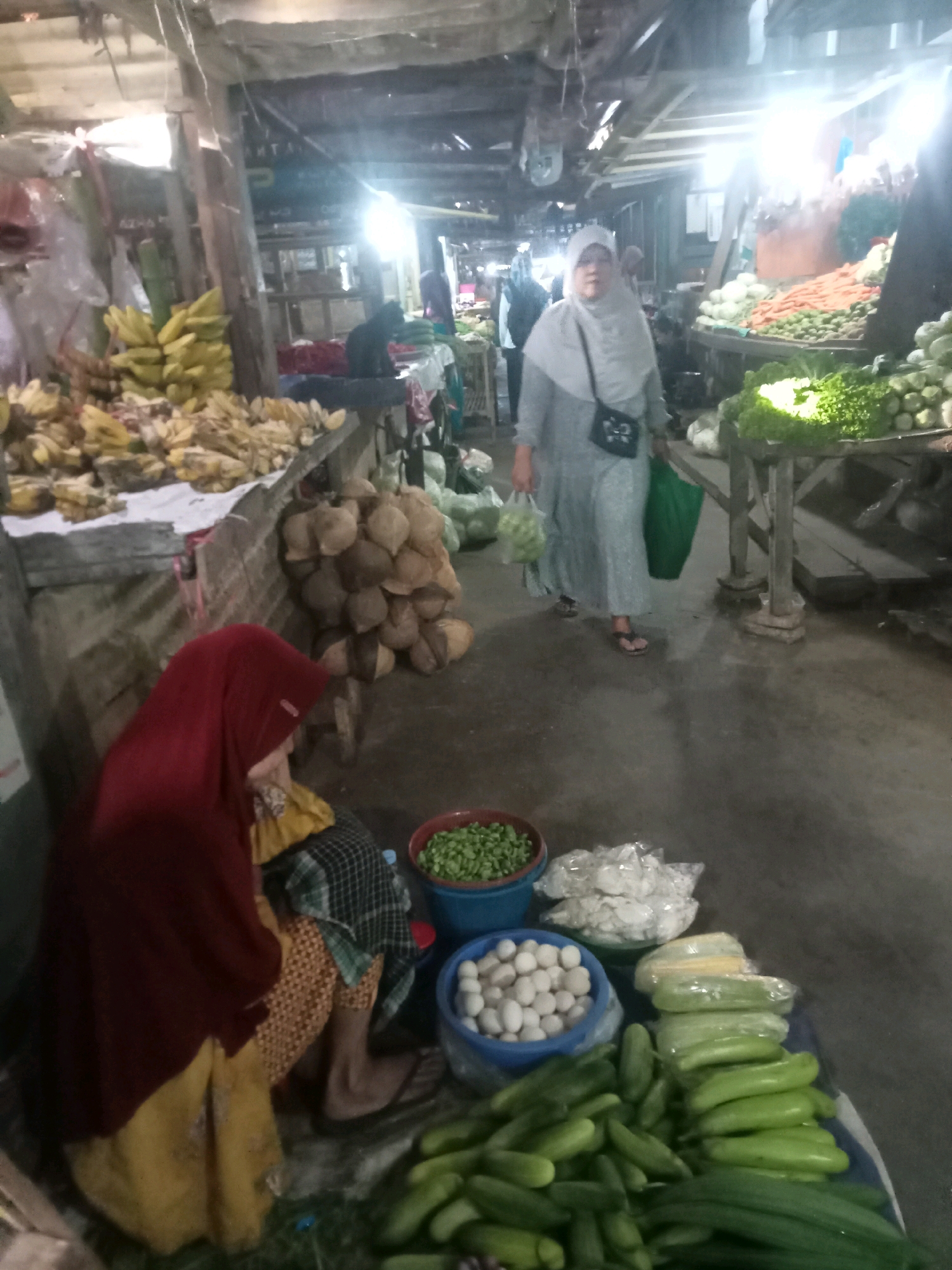 This is a full-shot, eye-level, interior photograph of a bustling market stall, likely in Probolinggo, Indonesia, given the presence of local produce and attire. The scene is dimly lit, with strong overhead lighting creating bright highlights and deep shadows. The stall is packed with a variety of fresh produce, including bananas, coconuts, various green vegetables like cucumbers and leafy greens, carrots, and what appear to be eggs in a blue bowl.
In the foreground, a woman wearing a red headscarf and a yellow and patterned dress is seated on the ground, facing away from the camera and towards the produce. To her left, a pile of coconuts is stacked. In the middle ground, a woman wearing a light grey hijab and a long grey dress walks away from the camera, carrying a green shopping bag in her left hand and a clear plastic bag filled with green produce in her right. She is wearing sandals. Behind her, other market-goers are visible in the distance, their figures blurred.
The stall is constructed from rustic wooden beams and planks, with corrugated metal or plastic sheeting forming the roof. The ground appears to be concrete or packed earth. The overall impression is one of a lively, local market selling fresh food.