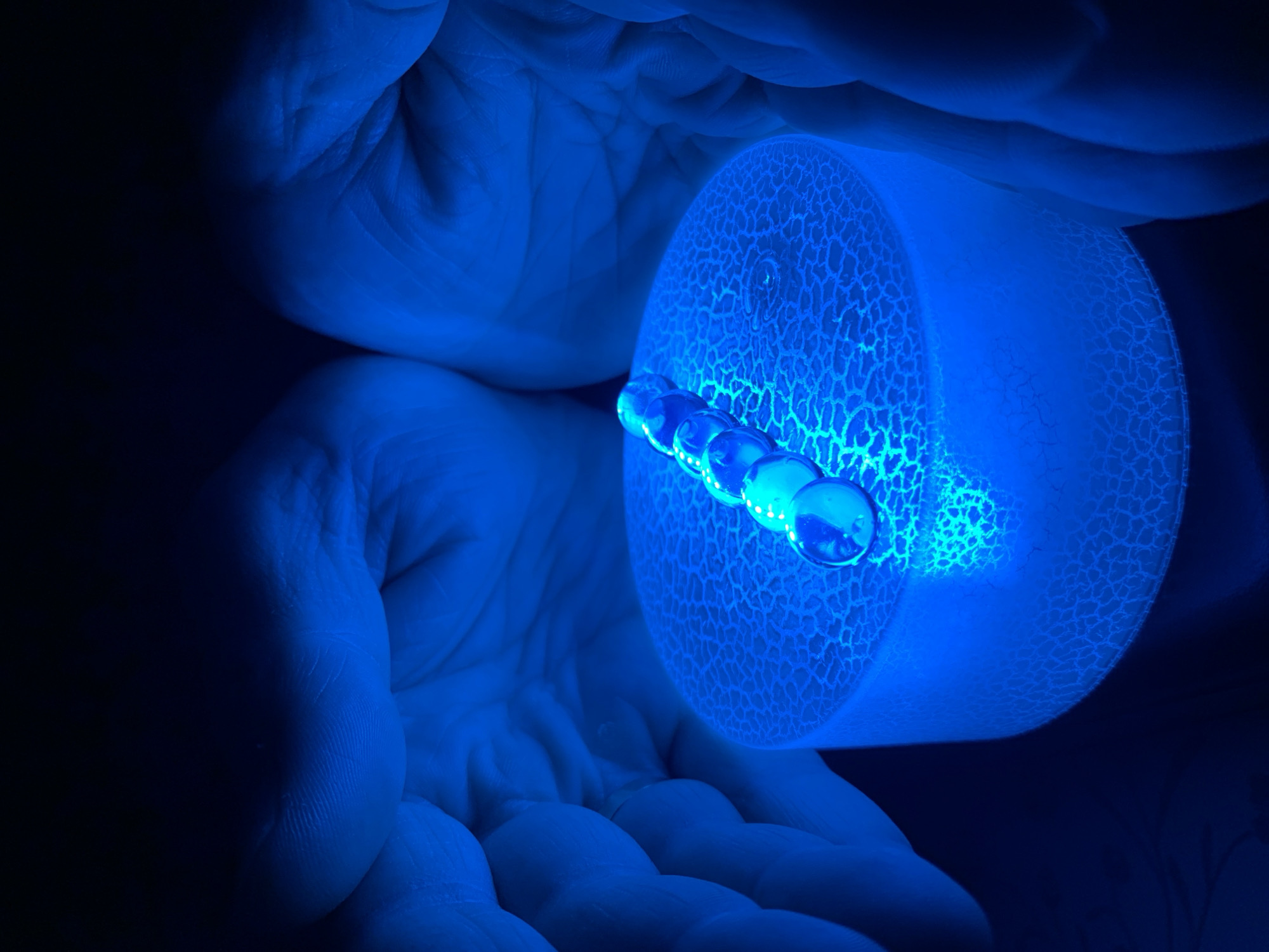 In a dimly lit, dark environment, a person's hands, illuminated by a vibrant blue light, cradle a round, cylindrical object. The object has a textured, cracked surface that glows intensely with the same blue light. Resting on top of this glowing cylinder is a line of clear glass marbles, some appearing to be swirled with white or blue patterns. The marbles are arranged in a linear fashion and are also reflecting the blue light. The overall impression is one of a mystical or scientific display, possibly under a black light or a colored LED light source. The image is focused on the marbles and the illuminated cylinder, with the hands providing a framing element. There is no indication of a specific location in Chaikovsky, Russia.