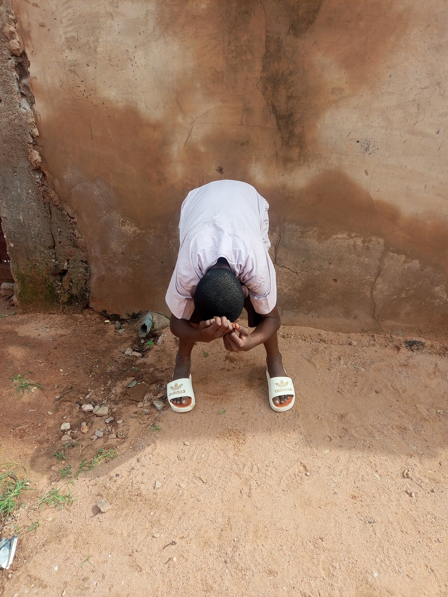 In the city of Tempure, Nigeria, a young Black male is pictured from behind, crouching and covering his face with his hands. He is wearing a light-colored traditional garment and white Adidas slides. The ground beneath him is sandy and uneven, with scattered small stones and a few blades of grass. Behind him is a textured, weathered, earth-toned wall that appears to be made of concrete or stucco. The wall has cracks and discolorations, suggesting age and exposure to the elements. A short, dark, cylindrical object protrudes from the wall near the ground, possibly a drainpipe. The lighting suggests it is daytime, with the sun casting some shadows. The overall scene is one of simplicity and potentially a moment of distress or contemplation.