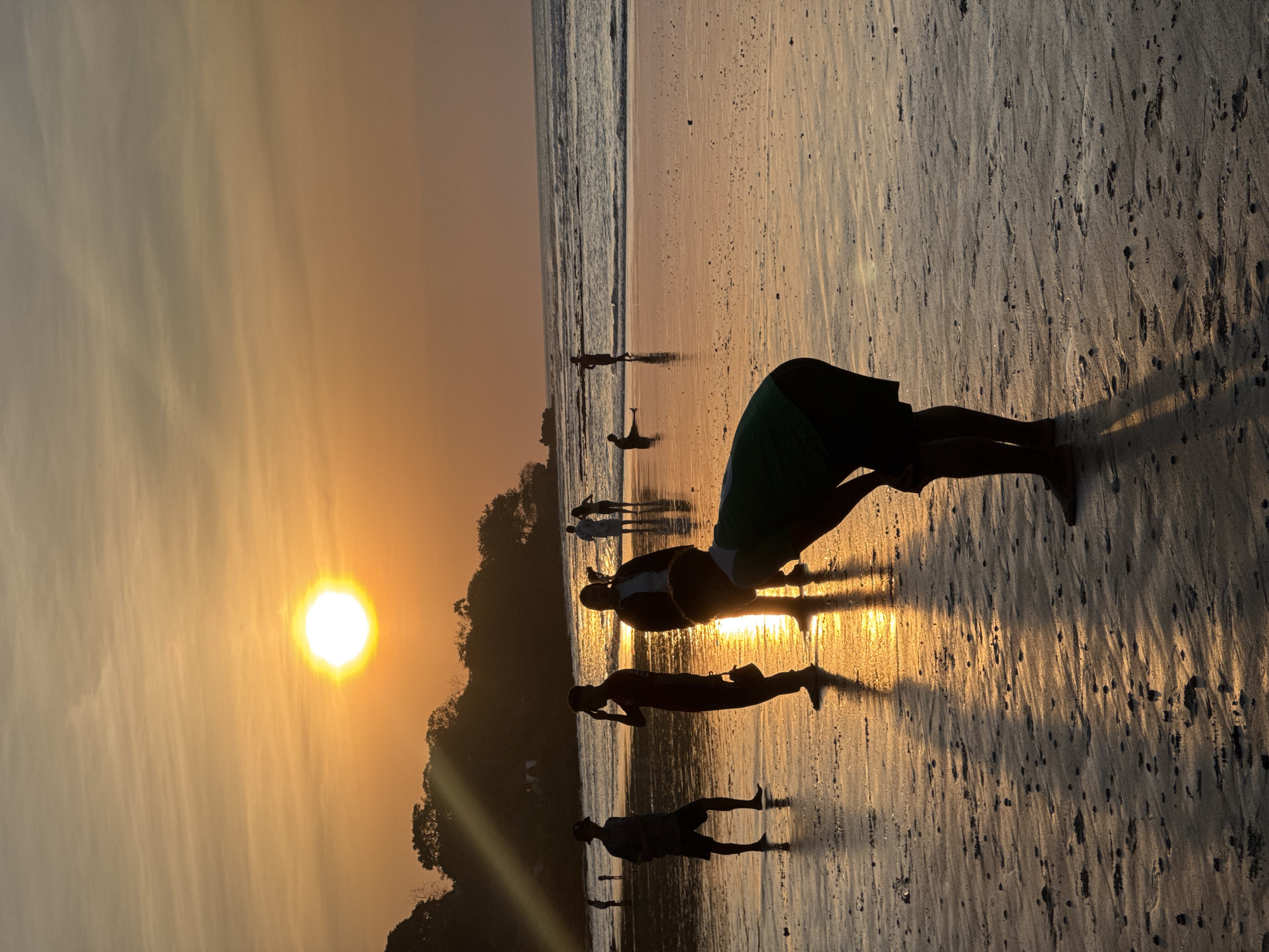 This image depicts a beach scene in Kuta Selatan, Indonesia, during sunset. The sun, a bright orb casting a warm orange glow, is positioned in the upper left quadrant of the sky, with thin clouds streaking across it. The foreground is a sandy beach wet from the receding tide, reflecting the golden light of the setting sun. Several people are scattered across the beach. In the middle ground, a group of individuals are silhouetted against the light. One person in the foreground, facing away from the camera, is bent over. Further back, the ocean waves gently lap the shore, with more silhouetted figures visible in and near the water. The background features a tree-covered hill or cliff. The overall atmosphere is serene and idyllic, characteristic of a tropical beach at dusk.