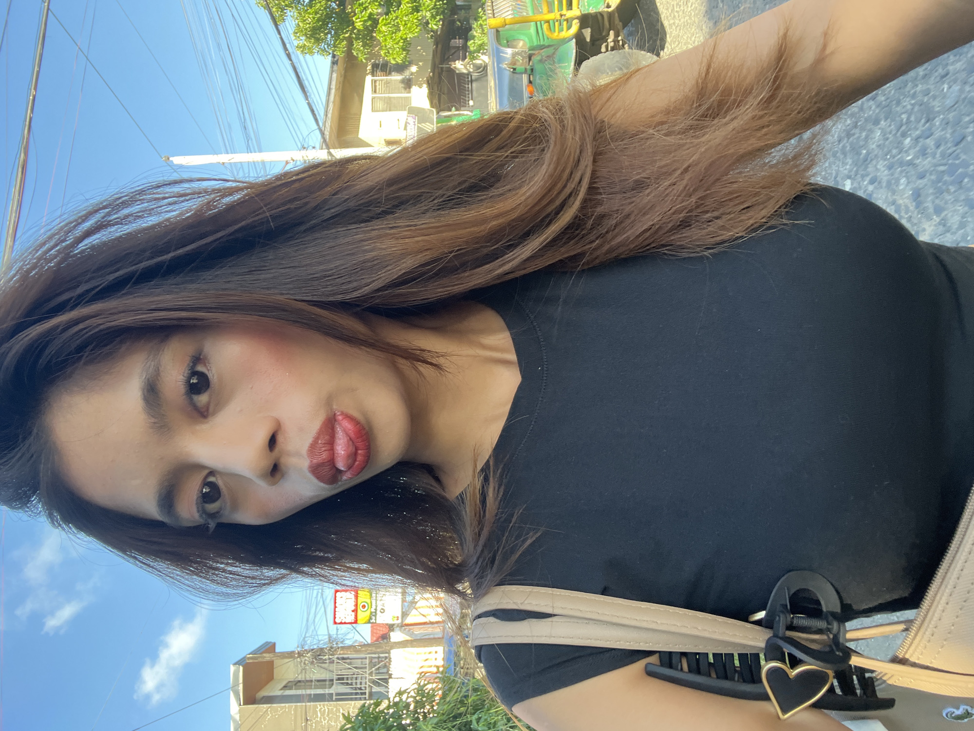 A woman with long, brown hair, wearing a black top and a beige bag, sticks her tongue out at the camera while standing on a street. She has on makeup, including red lipstick. A green vehicle is parked behind her, and there are buildings and power lines in the background. The scene appears to be in Davao City, Philippines, given the distinctive "MAMA" sign in the background, which is a known landmark in Davao City.