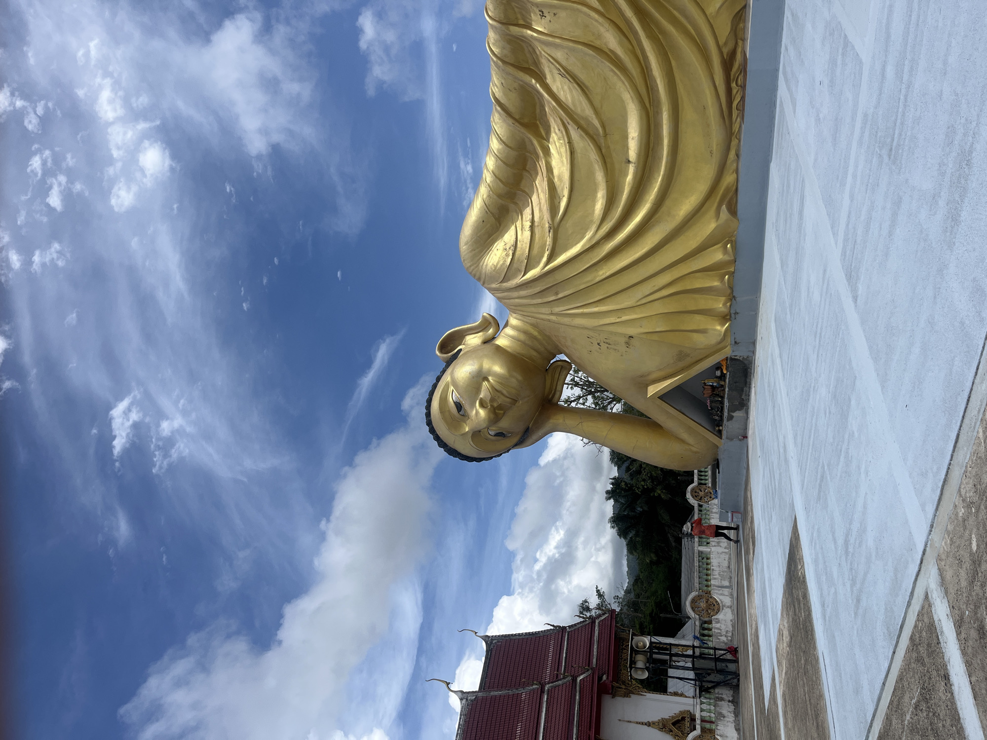 In the city of Si Sunthon, Thailand, a large, golden, reclining Buddha statue rests outdoors. The statue is golden and detailed, with the Buddha's head resting on its hand and arm. The Buddha is adorned in flowing robes, with prominent folds and creases. The statue is situated on a light gray platform, which is adjacent to a paved area that is being painted with what appears to be a light gray substance. A person wearing an orange shirt and a hat is seen painting the ground with a roller. To the left of the statue is a traditional Thai temple building with a red, tiered roof and white walls, decorated with ornate golden trim. The temple has a speaker system mounted on a stand. In the background, lush green trees and rolling hills are visible under a blue sky filled with scattered white clouds. The scene appears to be a religious site under development or maintenance.