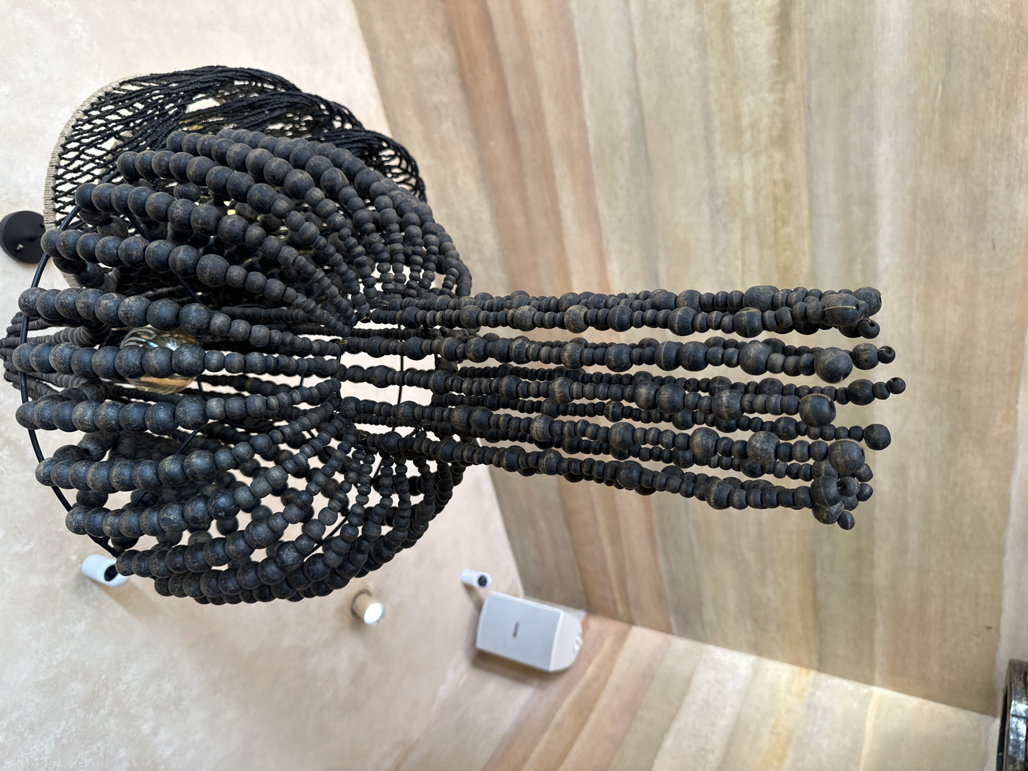 A dark beaded chandelier hangs from a textured ceiling. The chandelier features multiple strands of dark, round beads of varying sizes, creating a layered and cascading effect. A portion of the chandelier's top structure is visible, revealing a woven black material. Two small white security cameras and a white speaker are mounted on a wall to the left of the chandelier, which is set against a background of striped, earthy-toned walls. The scene is lit by an overhead light fixture. This image was taken in Kuta Selatan, Indonesia.