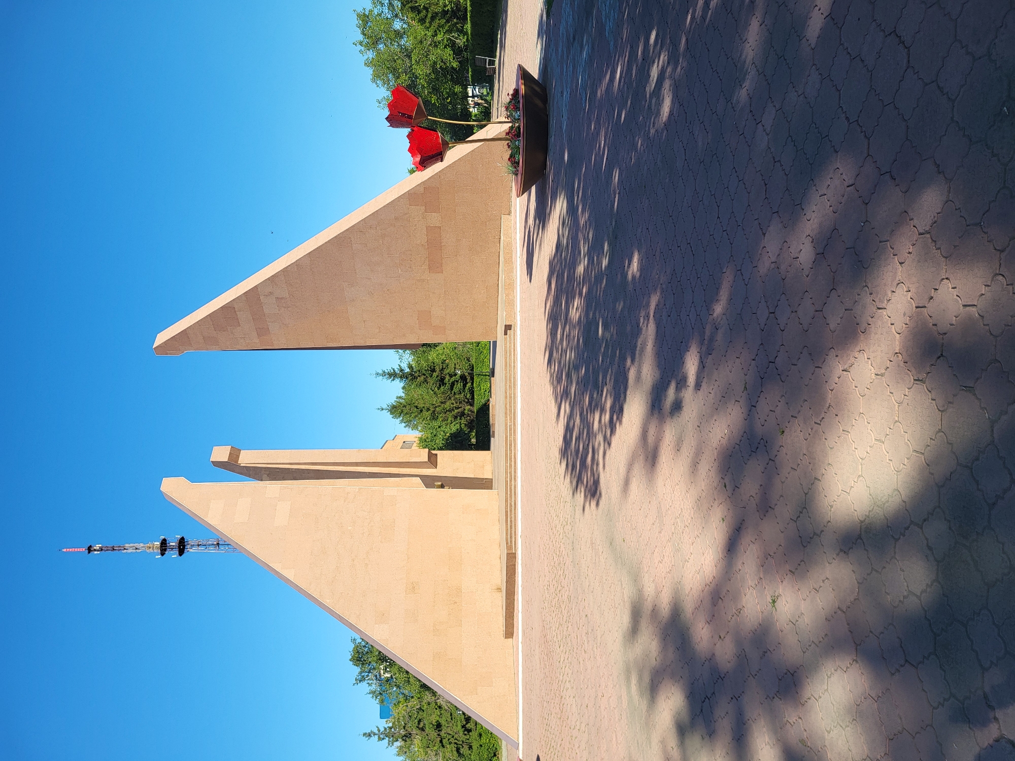 This image depicts a monument in the city of Petropavl, Kazakhstan. The monument consists of three large, angular beige stone slabs, two of which are significantly taller and lean inwards. In front of the monument, to the right, is a large, dark planter containing red floral decorations that resemble poppies. The ground in the foreground is paved with interlocking beige tiles, and dappled shade from trees falls across it. Behind the monument, evergreen trees are visible, and further back, a tall radio tower with a red and white antenna stands against a clear blue sky. The overall scene suggests an outdoor public space, possibly a park or memorial site.
