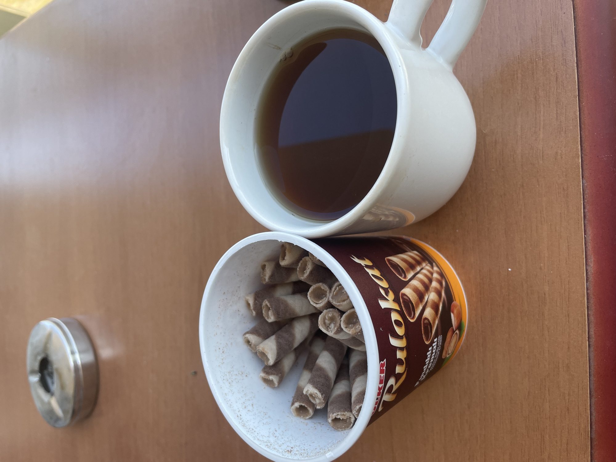 This media file depicts a close-up, overhead view of a wooden table with various items. The primary subjects are a white ceramic mug filled with dark liquid, likely tea or coffee, and a container of wafer roll snacks. The mug is positioned on the upper right side of the frame, and the container of snacks is below and to the left of the mug. The container appears to be made of cardboard and is adorned with branding that includes images of the wafer rolls and text. Some of the wafer rolls are visible spilling out of the container. To the far left of the frame, a small, circular metal object, possibly an ashtray or a small tin, is present. The lighting suggests it is daytime, with natural light illuminating the scene. The overall impression is of a casual break or snack time, likely indoors or in a sheltered outdoor area given the presence of the mug and snacks. There is no visible text that is legible for identification. The setting is a simple wooden surface, with hints of a darker, possibly red border visible on the right edge of the frame. The scene is static, with no discernible activity beyond the arrangement of the objects.