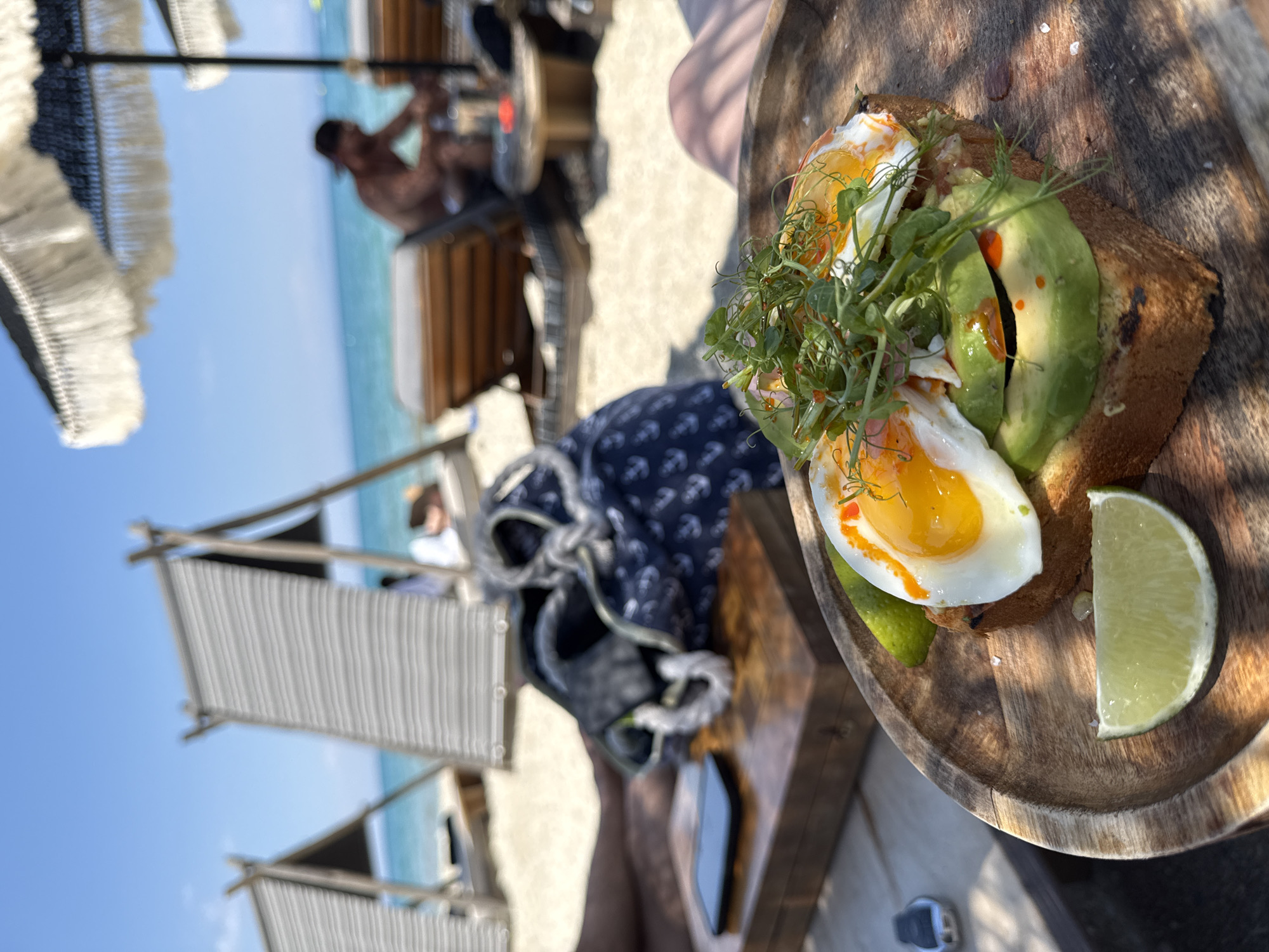 A close-up shot focuses on an avocado toast with two fried eggs and microgreens, served on a wooden plate with a lime wedge on a wooden table. In the background, a beach scene unfolds with lounge chairs, umbrellas, and the turquoise sea. A person can be seen relaxing on a lounge chair, partially obscured by an umbrella. The image was taken in the Municipal Unit of Pallini, Greece.