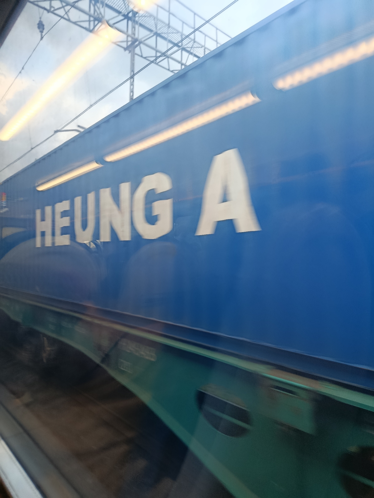 The image shows a moving train car with the words "HEUNG A" written in large white letters on its side. The train car is blue and has a teal-colored bottom section. There are bright yellow horizontal lights visible on the side of the train car and also reflected in the sky. Overhead, there are power lines and metal structures, suggesting the train is on an electrified railway line. The image appears to be taken from another moving vehicle, likely a train, looking out a window, as evidenced by the motion blur and reflections. The environment is a mix of industrial infrastructure and a cloudy sky. The exact location in the city of Elektrougli, Russia, cannot be precisely determined from the visual information alone, but the context suggests a railway environment within or near the city.