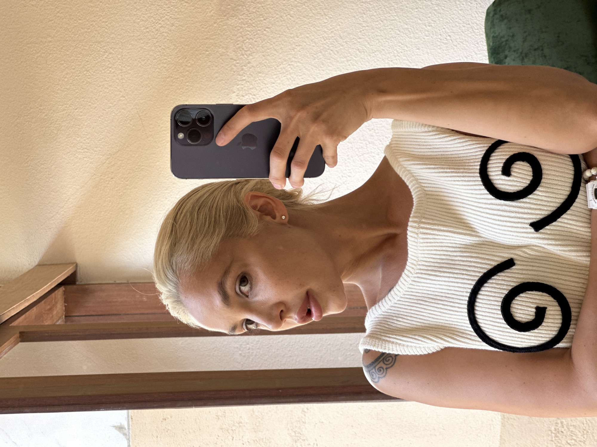 A woman is taking a selfie indoors in what appears to be Kuta Selatan, Indonesia. She is holding a dark-colored smartphone in her right hand, positioning it above her head. The woman has light blonde hair, and is wearing a white ribbed top with black spiral designs. She has a tattoo on her upper left arm. The background includes a beige wall and a set of wooden stairs. The lighting is bright, suggesting daylight. A green object is visible in the top right corner.