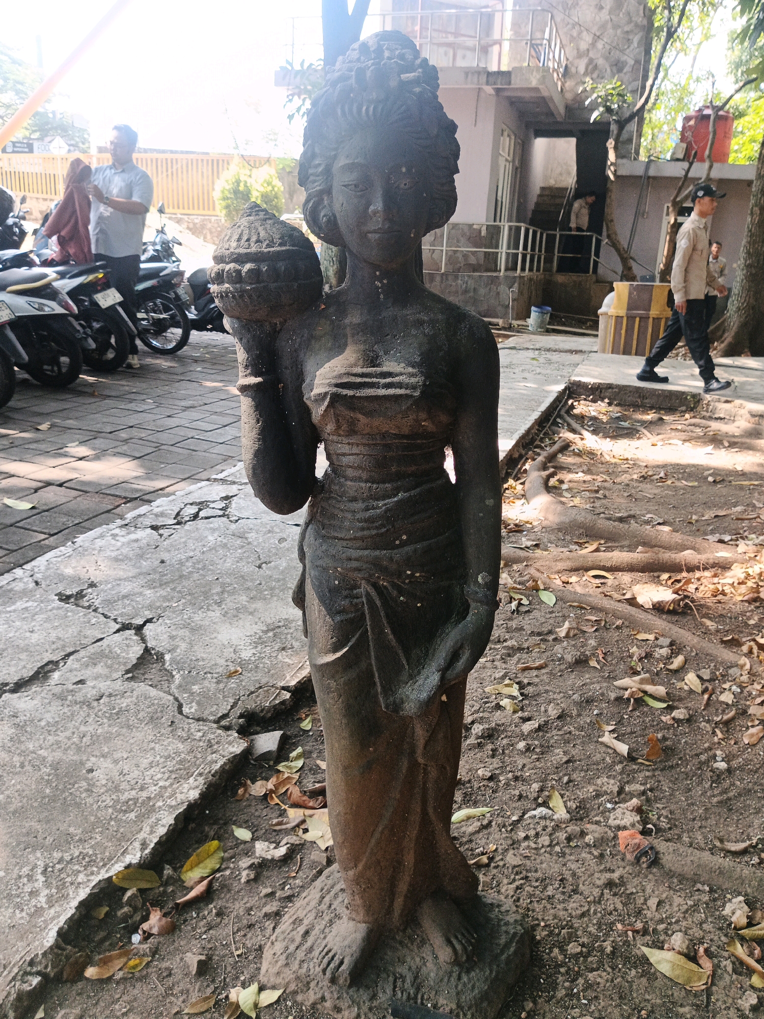 The image shows a stone statue of a woman in the foreground, holding a large object on her left shoulder. She is dressed in traditional clothing. In the background, several motorcycles are parked on a tiled ground. A man in a light-colored shirt and dark pants is standing near the motorcycles, holding a maroon garment. Further in the background, a building with multiple stories is visible, with stairs leading up to it. Two men in light-colored uniforms are walking on a paved path. The environment appears to be an outdoor area with trees and fallen leaves on the ground. The scene is illuminated by natural daylight, creating shadows. The location is identified as the city of Bandung, Indonesia.
