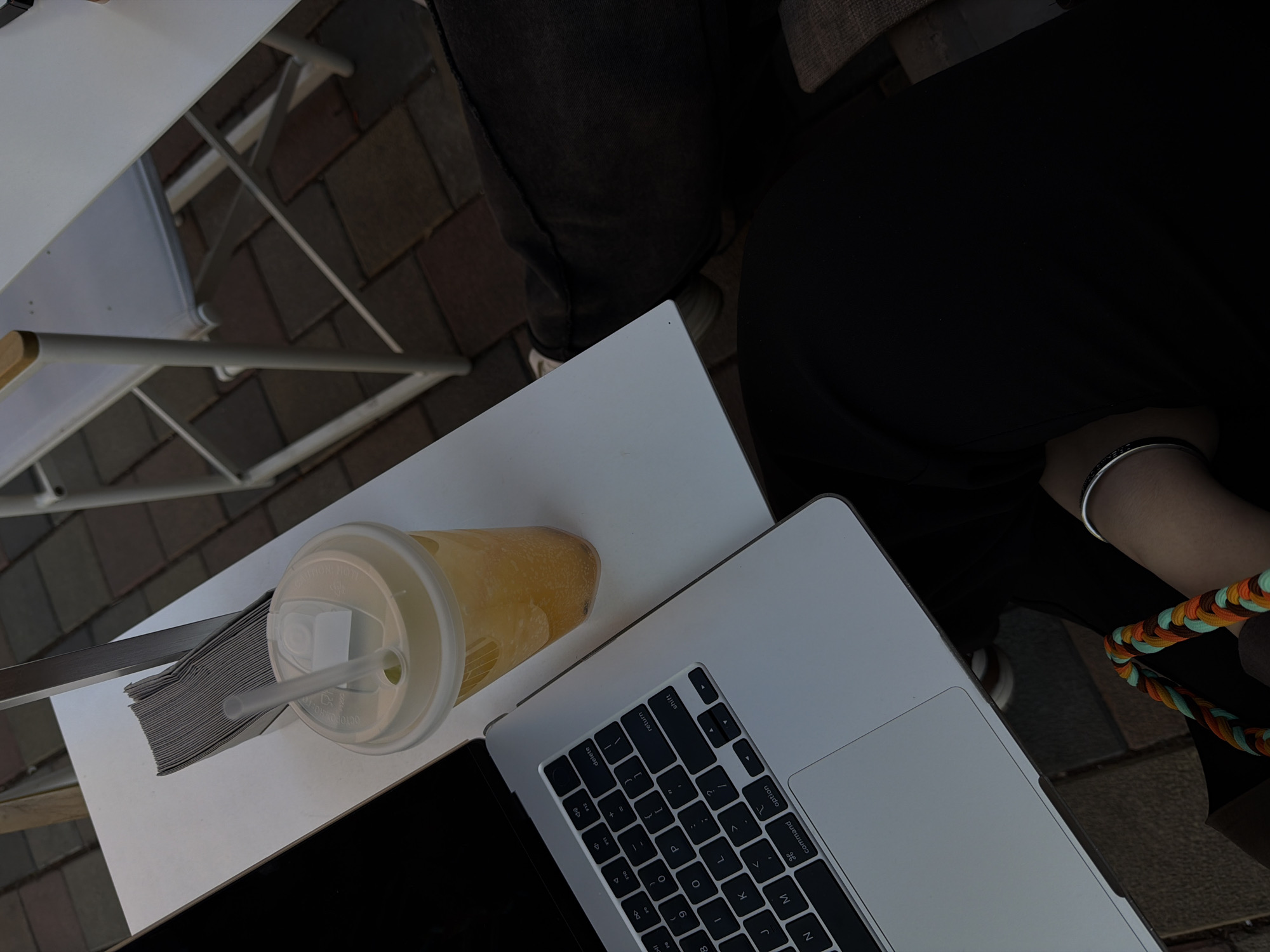 A high-angle shot depicts a person seated outdoors at a white table with a laptop, a beverage in a plastic cup, and a stack of napkins. The laptop is open and partially obscuring the beverage. The person is wearing dark clothing and a silver bracelet on their wrist, with a colorful braided strap visible near their hand. The table appears to be set on a brick or tiled surface. Given the urban setting and the presence of a laptop and a beverage, this scene might be taking place in a cafe or outdoor workspace. However, the location cannot be confirmed to be Yerevan, Armenia, as there are no specific identifiers.